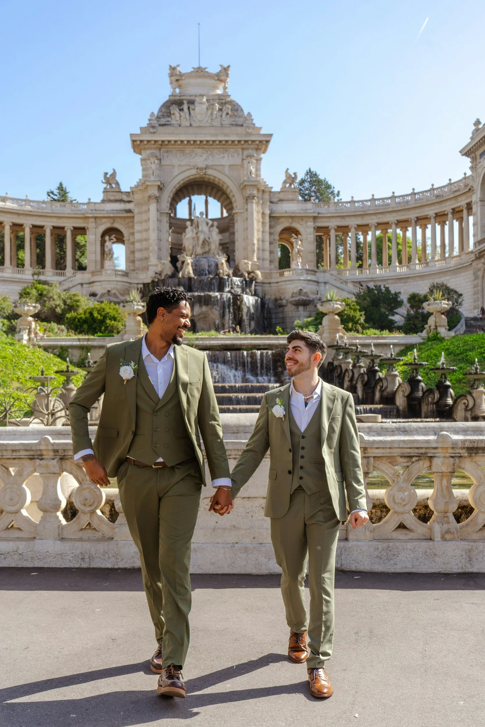 Two grooms in matching green suits holding hands and smiling in front of an ornate fountain.