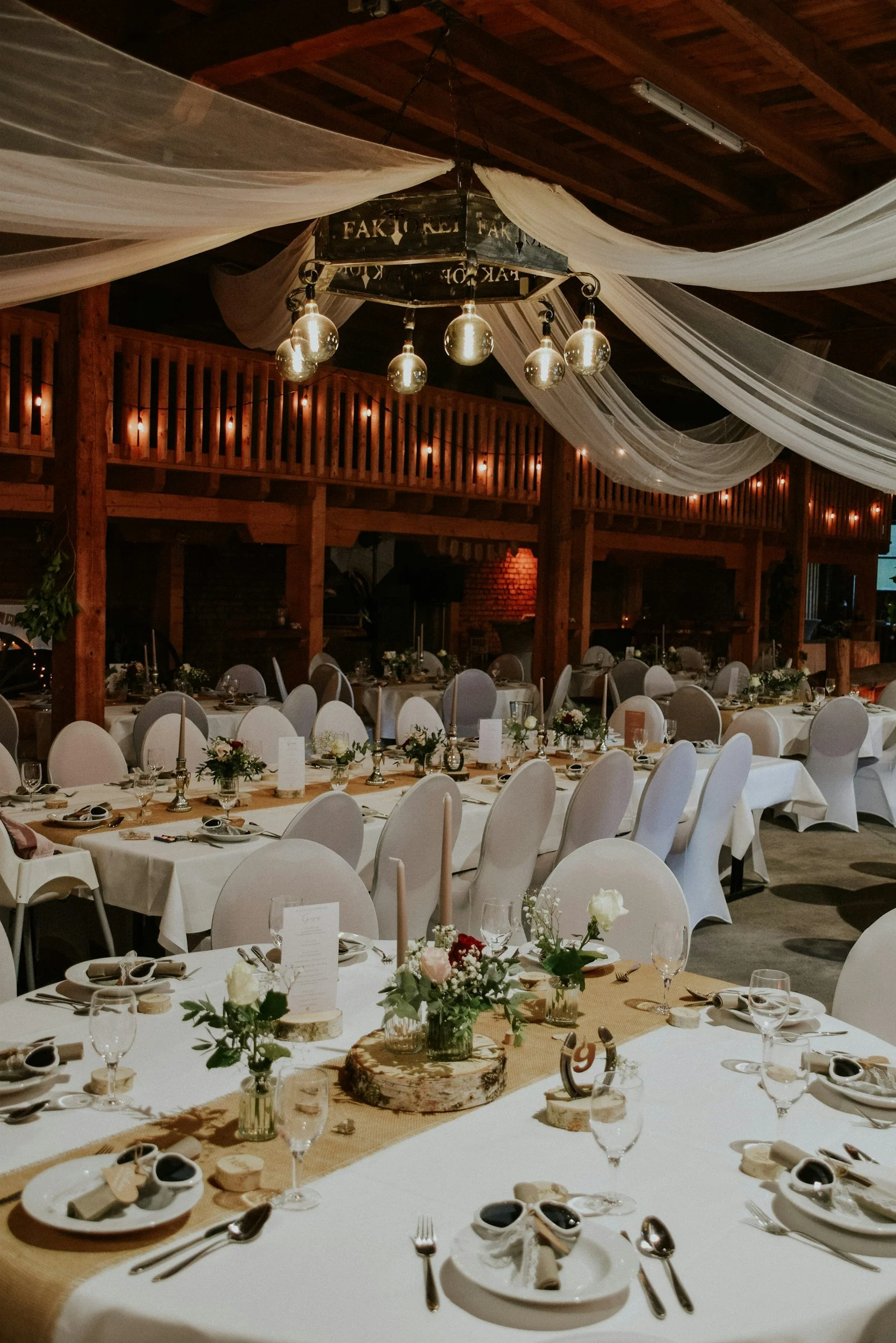 Rustic wedding reception with long tables, floral centerpieces, white draping, and hanging lights.