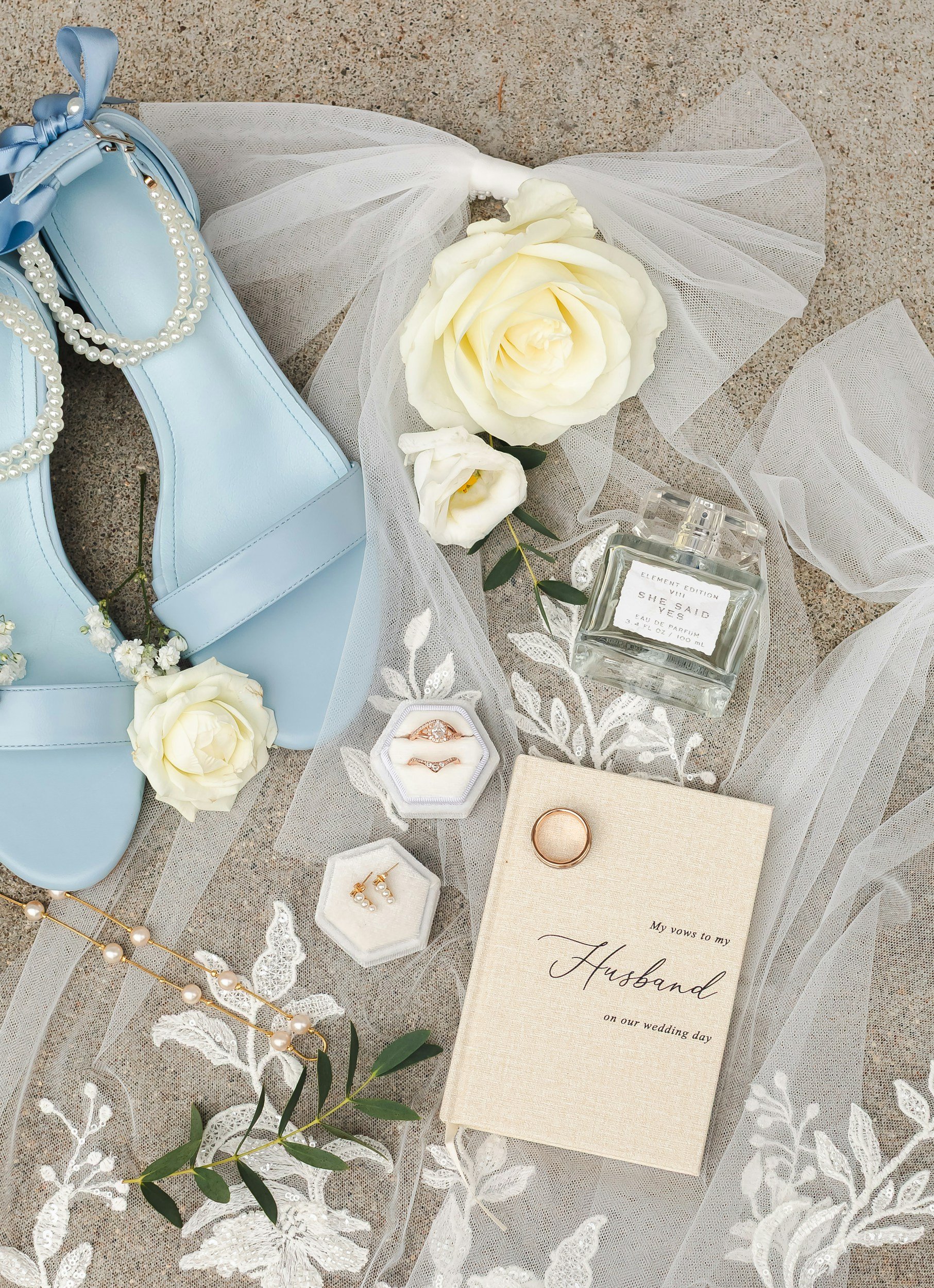 Bridal details including wedding rings, pearl earrings, perfume, white roses, veil, and a vow book.