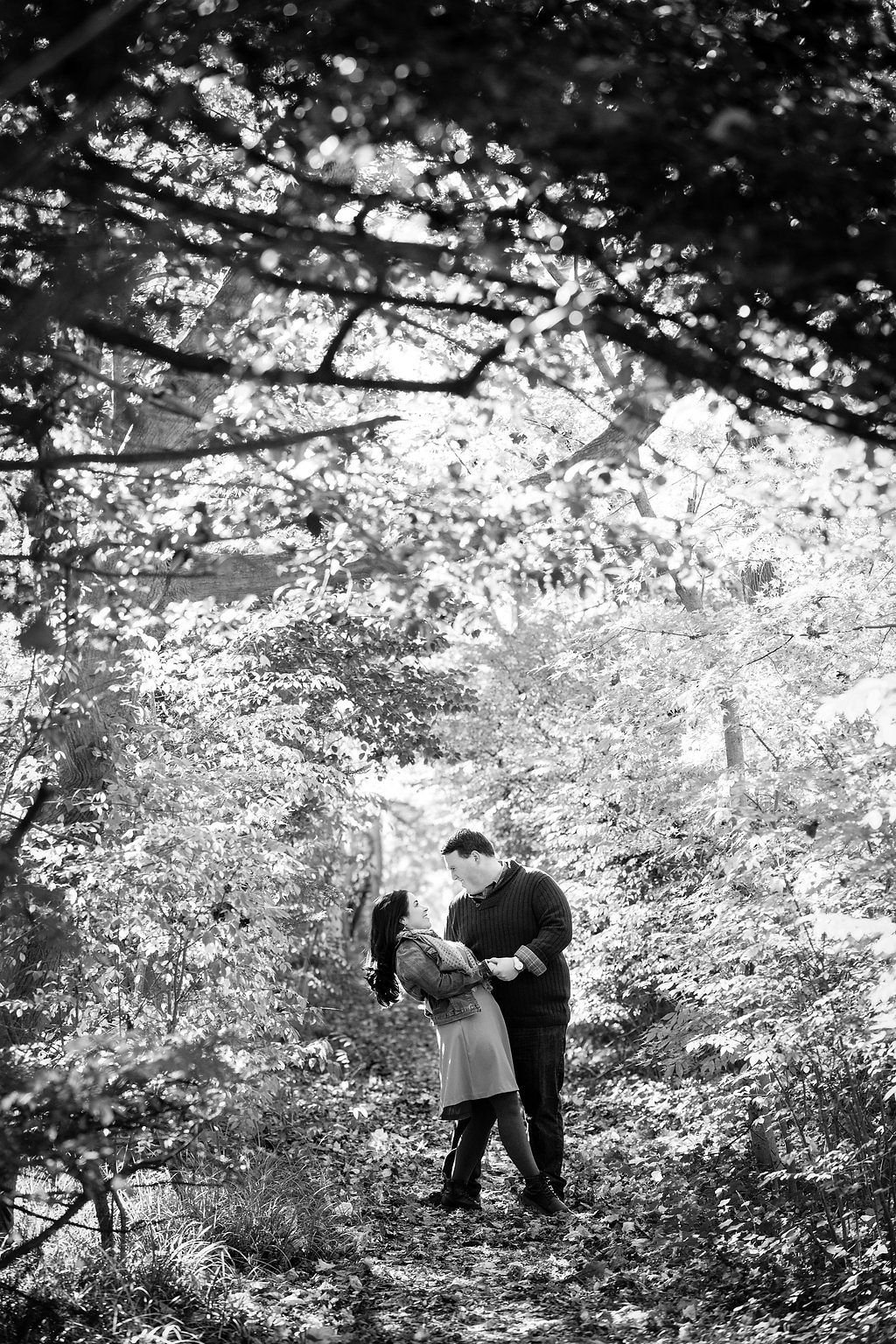 Black and white portrait of a couple embracing outdoors beneath the trees.
