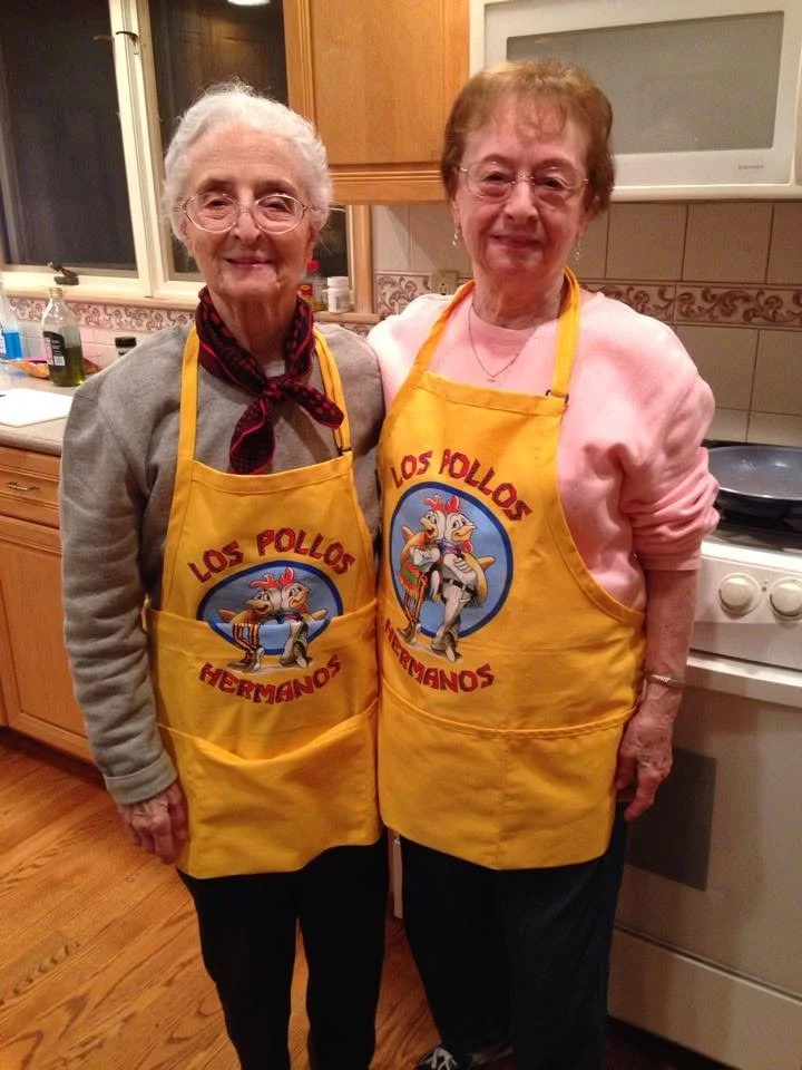 Two elderly women smiling together in a kitchen while wearing matching yellow Los Pollos Hermanos aprons.