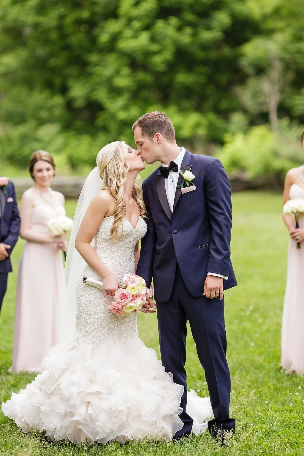 Bride and groom kissing outdoors while holding hands, with a bridesmaid in the background.