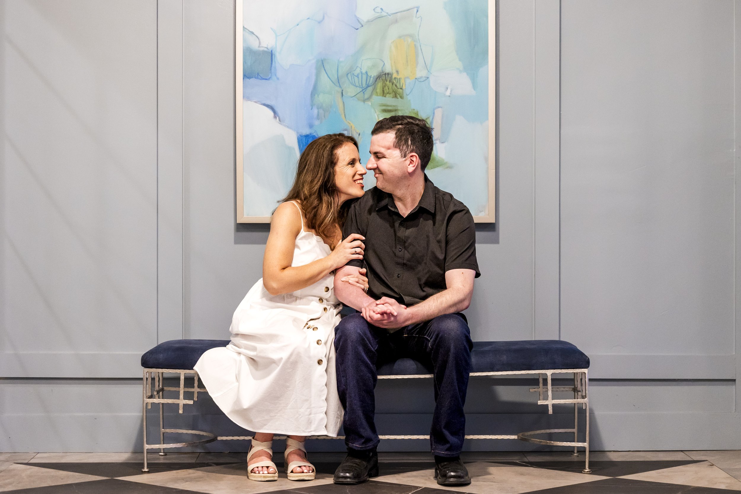 Couple sitting together and smiling at each other during an indoor engagement photo session.