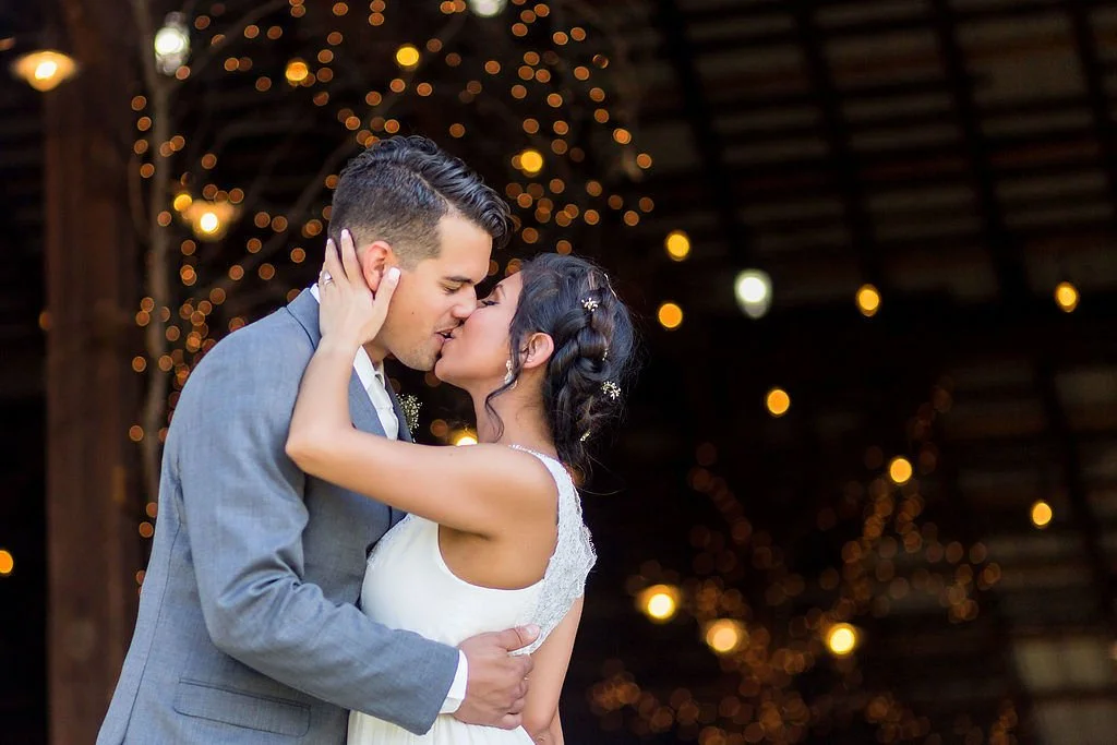 Bride and groom kissing during their wedding celebration with warm lights in the background.