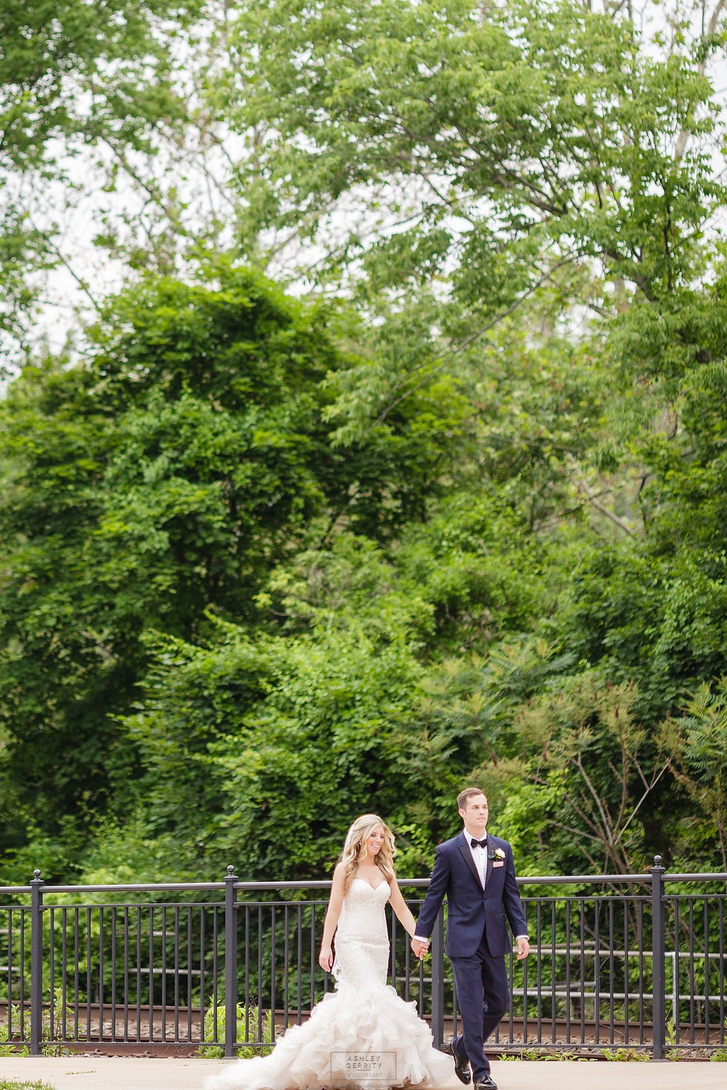 Bride and groom walking hand in hand across a bridge surrounded by lush greenery.