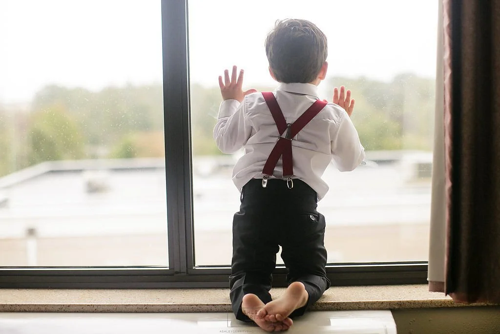Young boy in suspenders kneeling by a large window and looking outside.