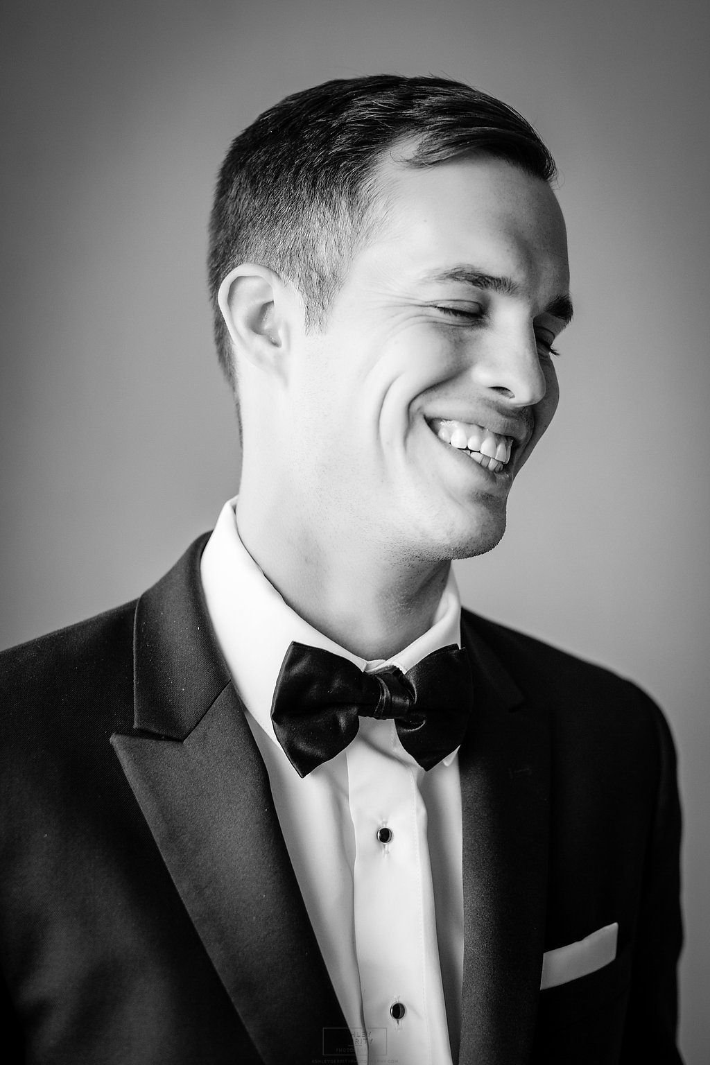 Black and white portrait of a smiling groom wearing a tuxedo and bow tie.