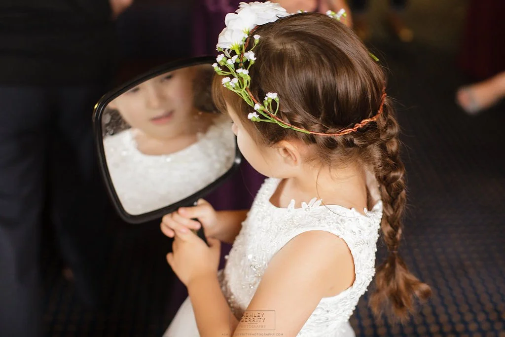 Young flower girl wearing a floral crown and looking at her reflection in a mirror.