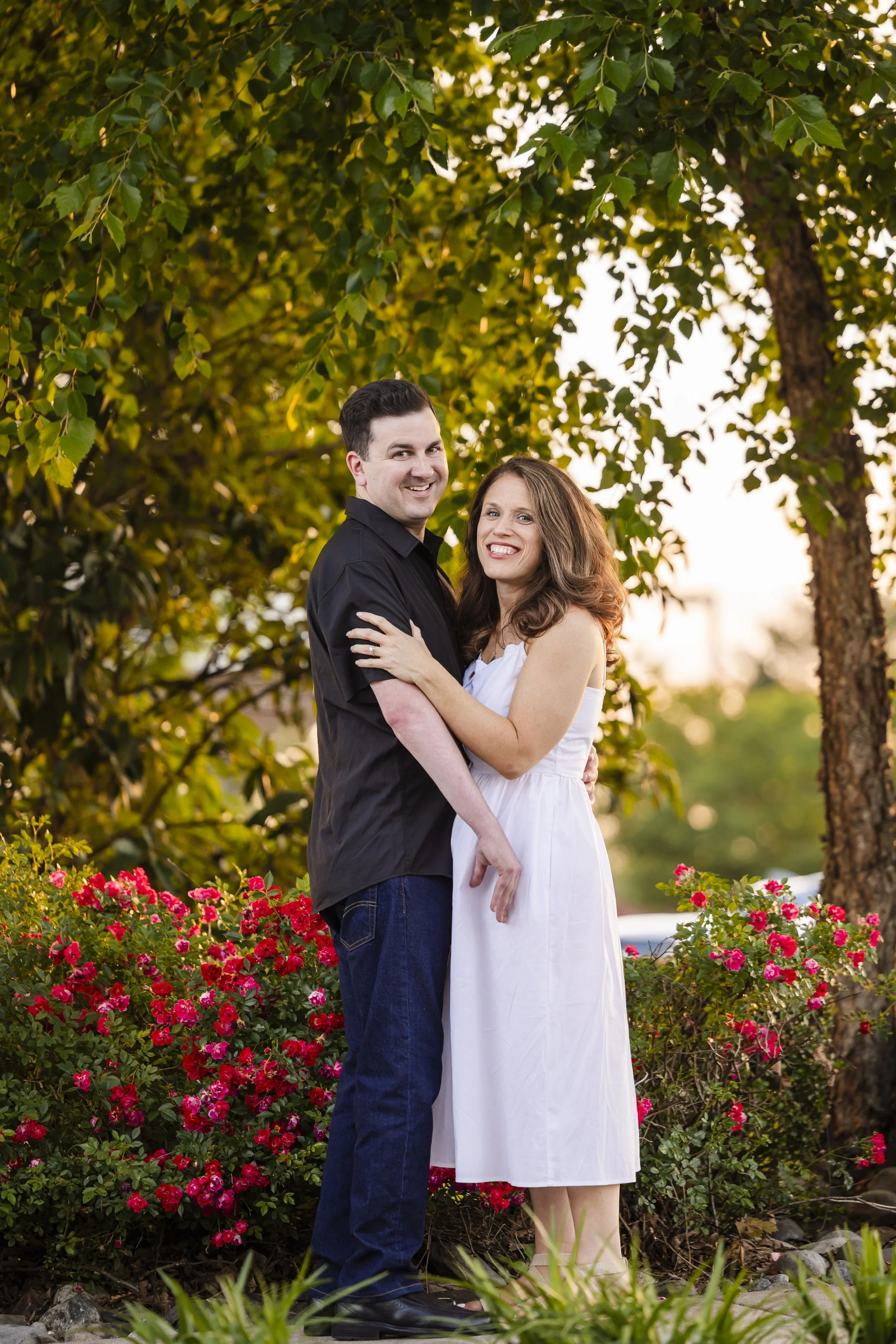 Engaged couple posing together outdoors in a garden with red flowers.