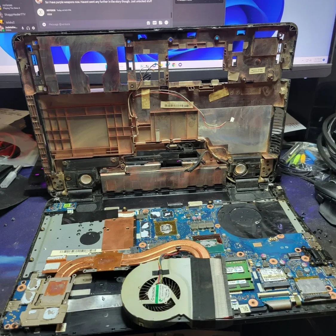 Open laptop with exposed internal components, including a cooling fan, copper heat pipes, and a blue circuit board, placed on a cluttered desk in front of a monitor.