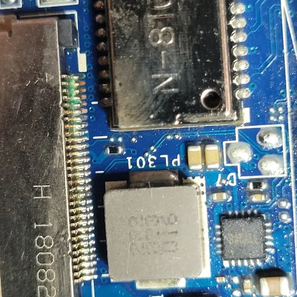 Close-up of a blue circuit board with various electronic components, including a metal shielded chip, a black socket, and multiple small capacitors and resistors.