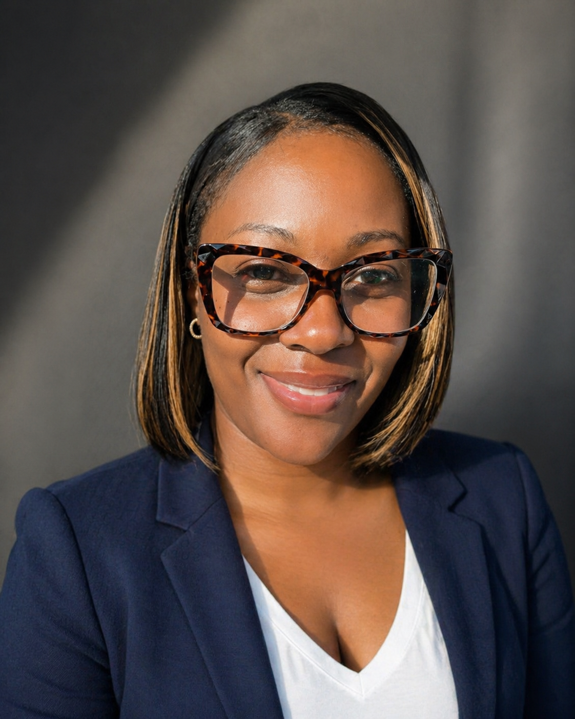 A woman with shoulder-length hair wearing large tortoise-shell glasses, a navy blazer, and a white top, smiling at the camera against a gray background.