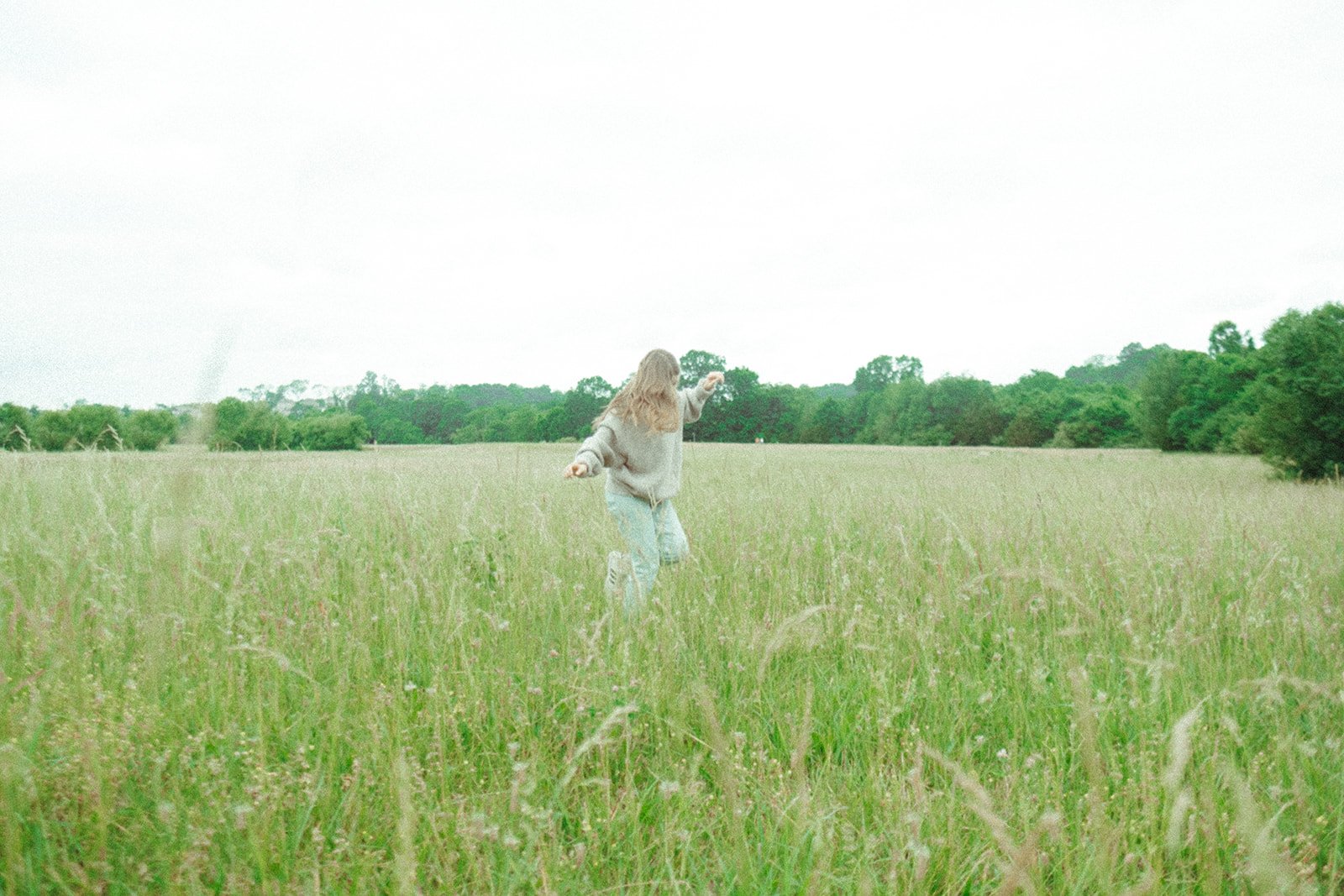 A person in a gray sweatshirt and jeans walking through a grassy field with trees in the background on a cloudy day.