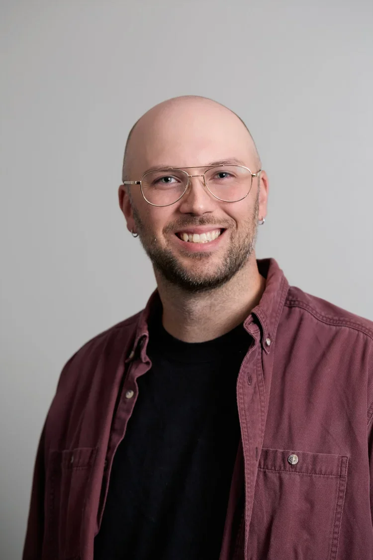 A smiling man with glasses, earrings, and a beard, wearing a maroon button-up shirt over a black t-shirt, photographed against a plain light gray background.