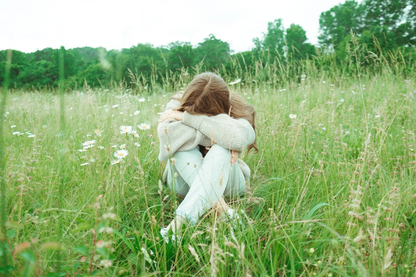 A person sitting on grass in a field, hugging their knees with their head down, surrounded by daisies and tall green grass, with trees and an overcast sky in the background.