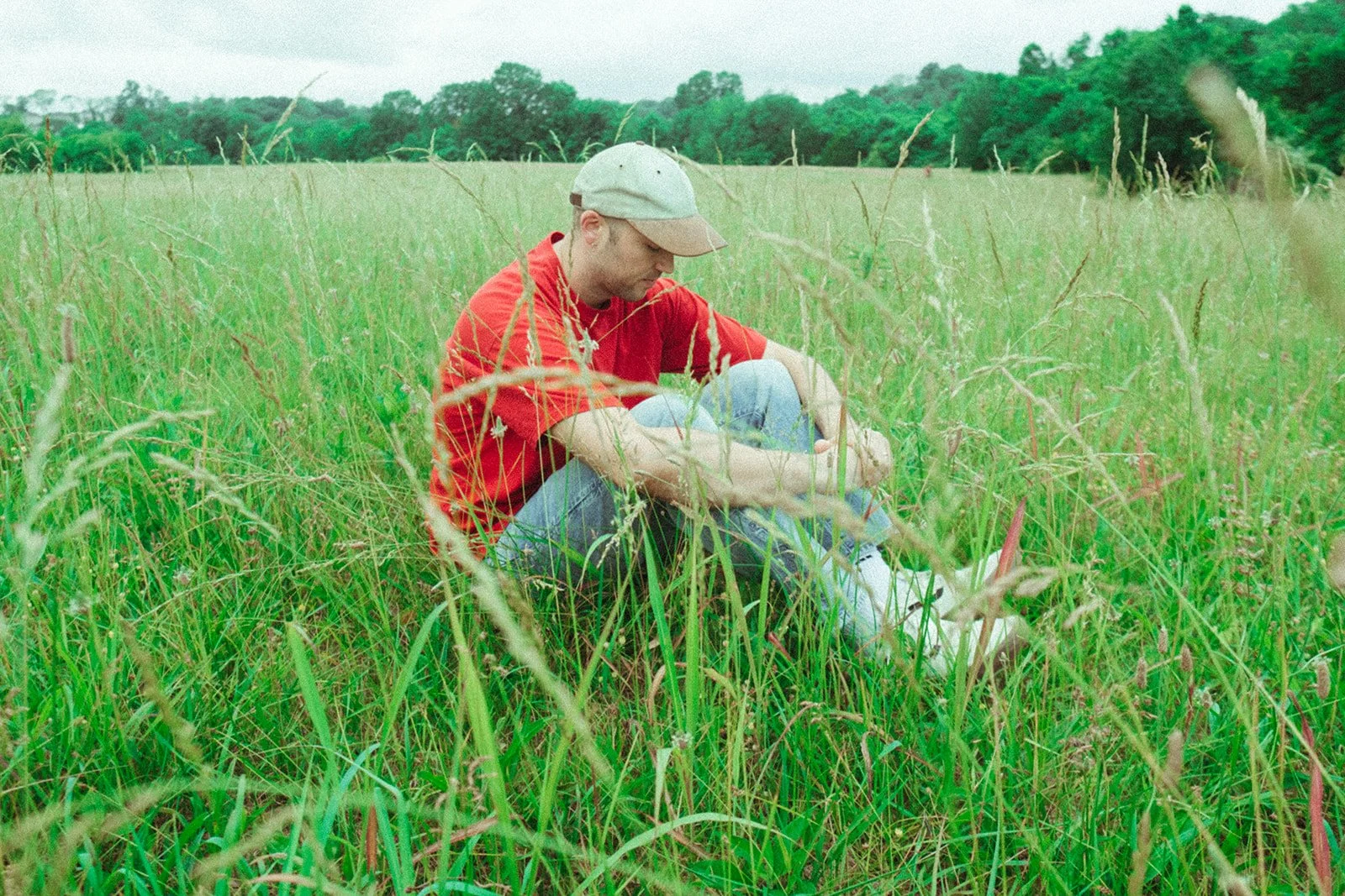 A man sitting in tall green grass in a field, wearing a red shirt and a gray baseball cap, looking downward
