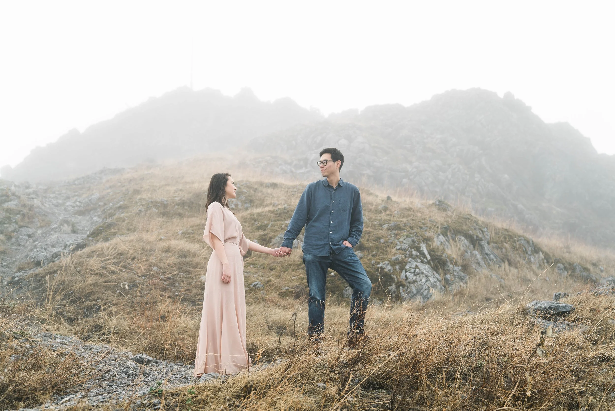 A couple holding hands outdoors on a grassy, rocky hillside under a misty, overcast sky, with a dark mountain ridge in the foggy background.