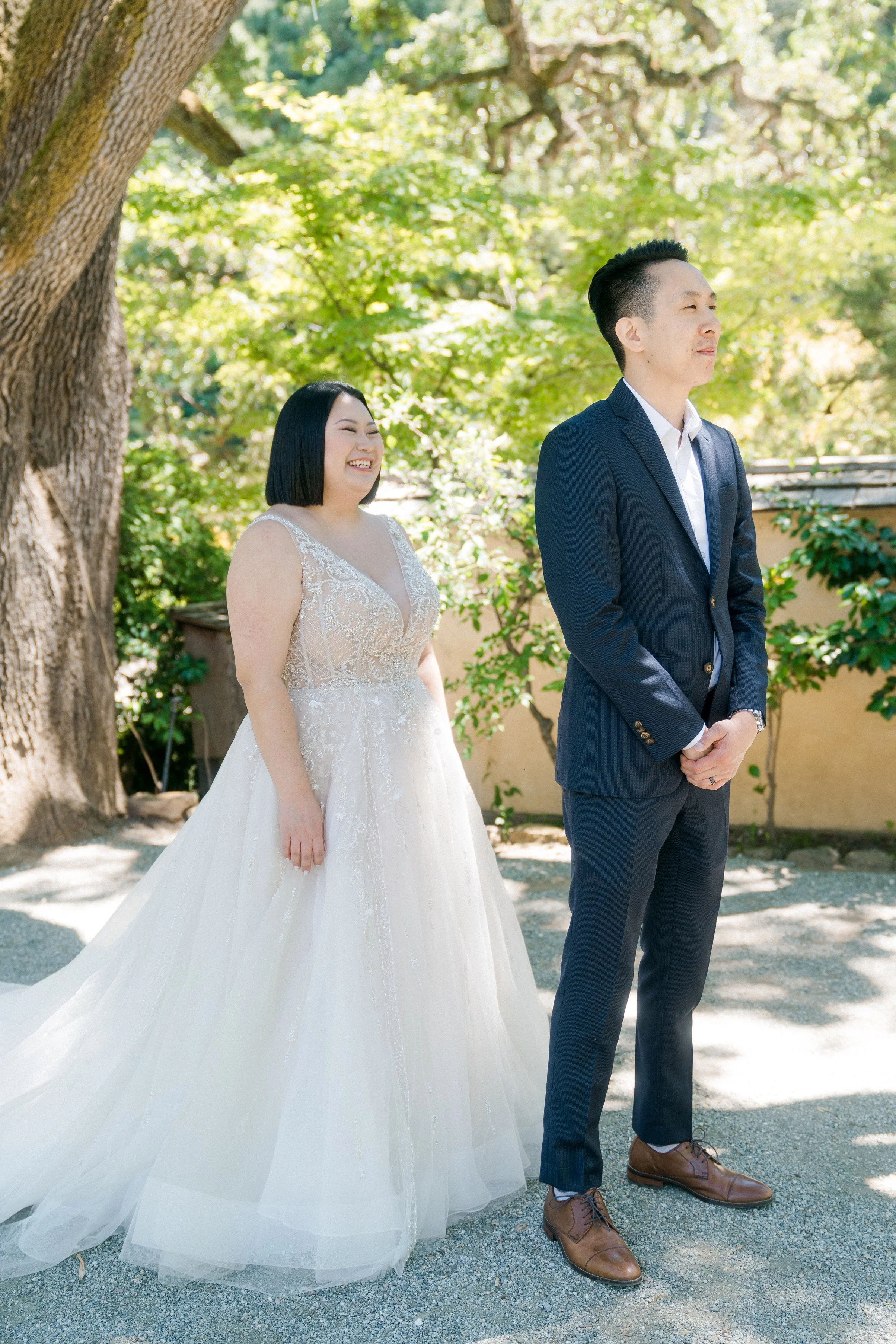 A smiling bride approaches her groom from behind for a wedding first look in the garden at Hakone Estate and Gardens in Saratoga, California.
