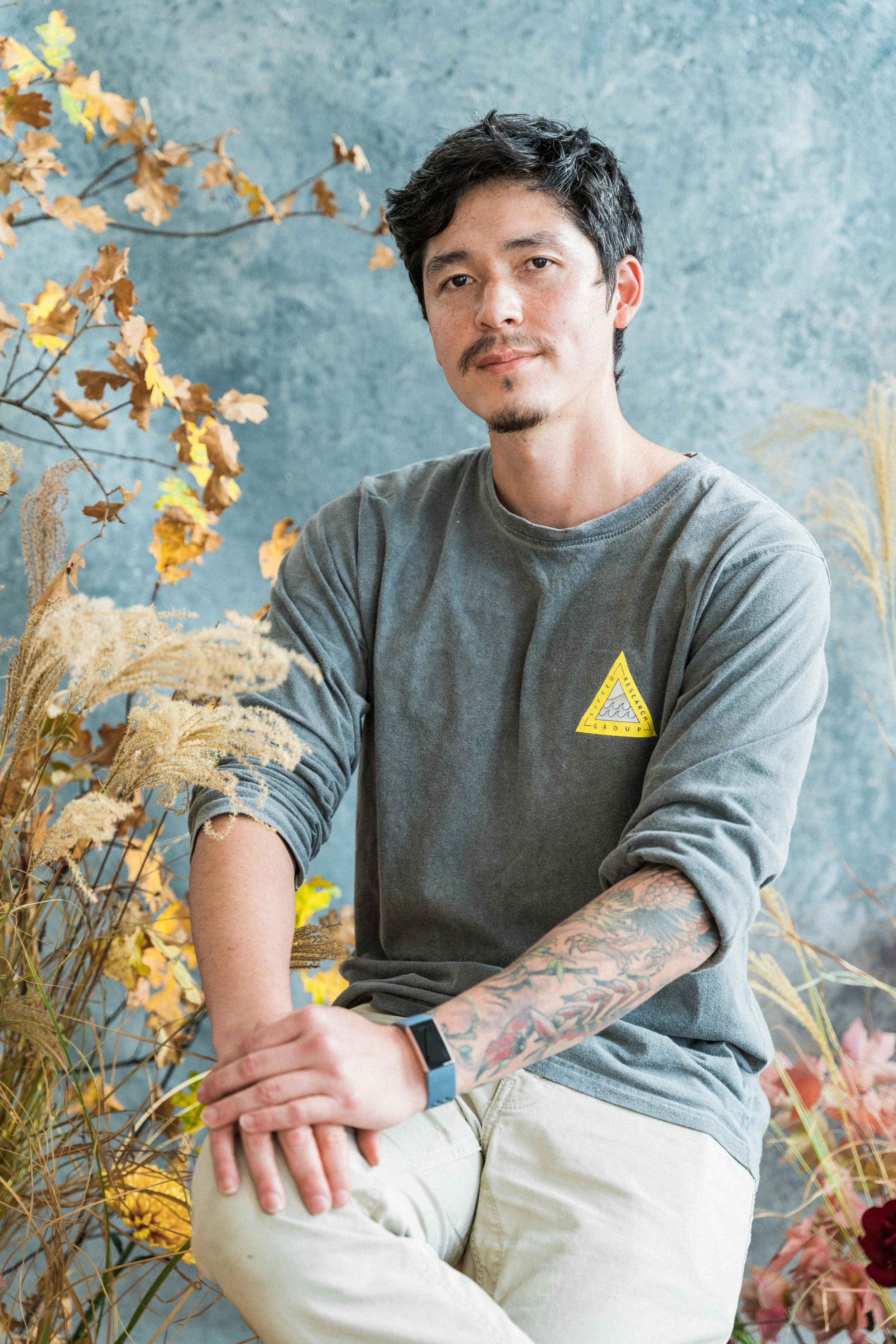 Portrait of Nico Lee-Quinones sitting comfortably against a textured blue backdrop surrounded by dried autumnal foliage.