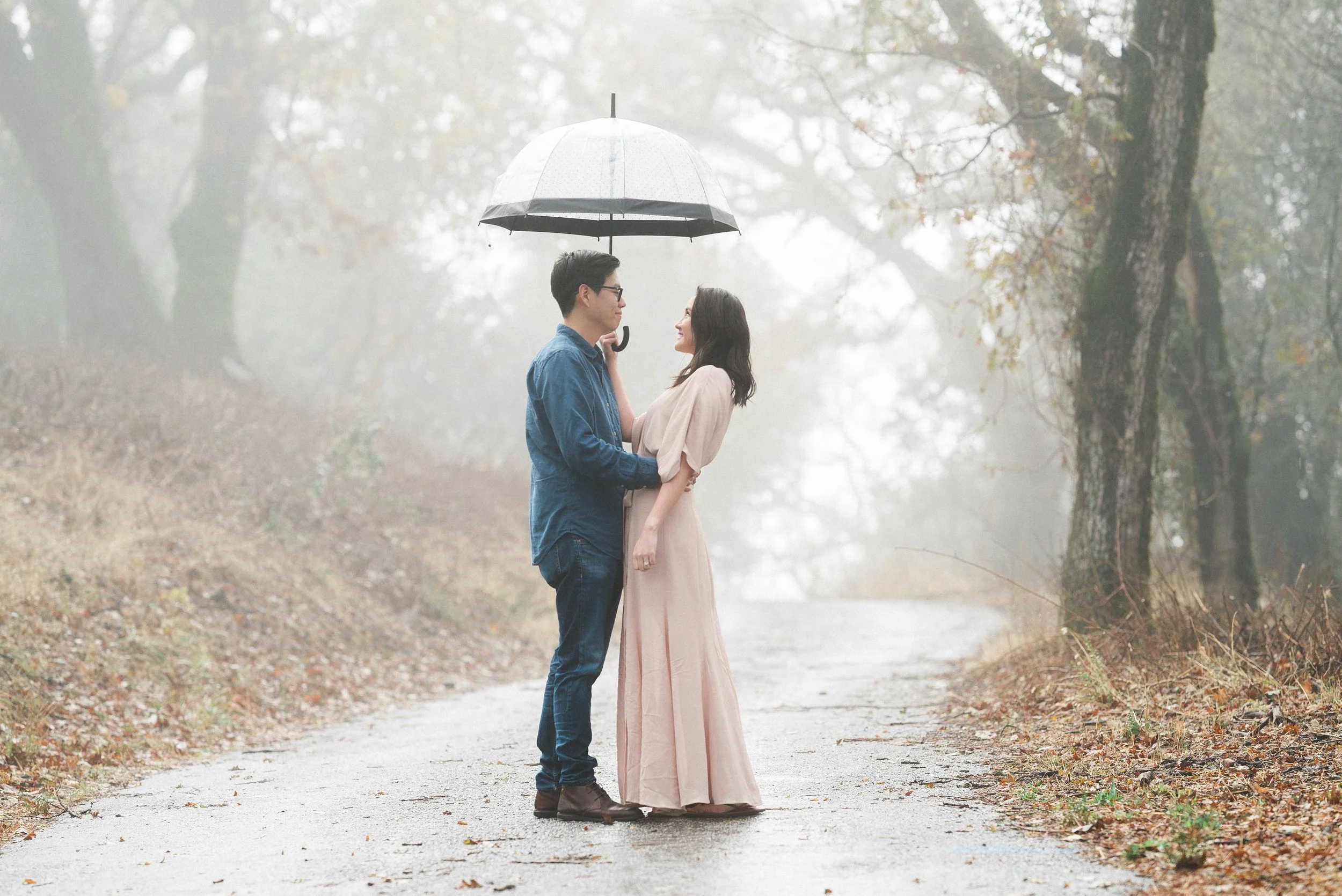 A man in a blue button-down shirt and a woman in a long blush pink dress holding a clear umbrella together while standing on a wet country road surrounded by foggy trees.
