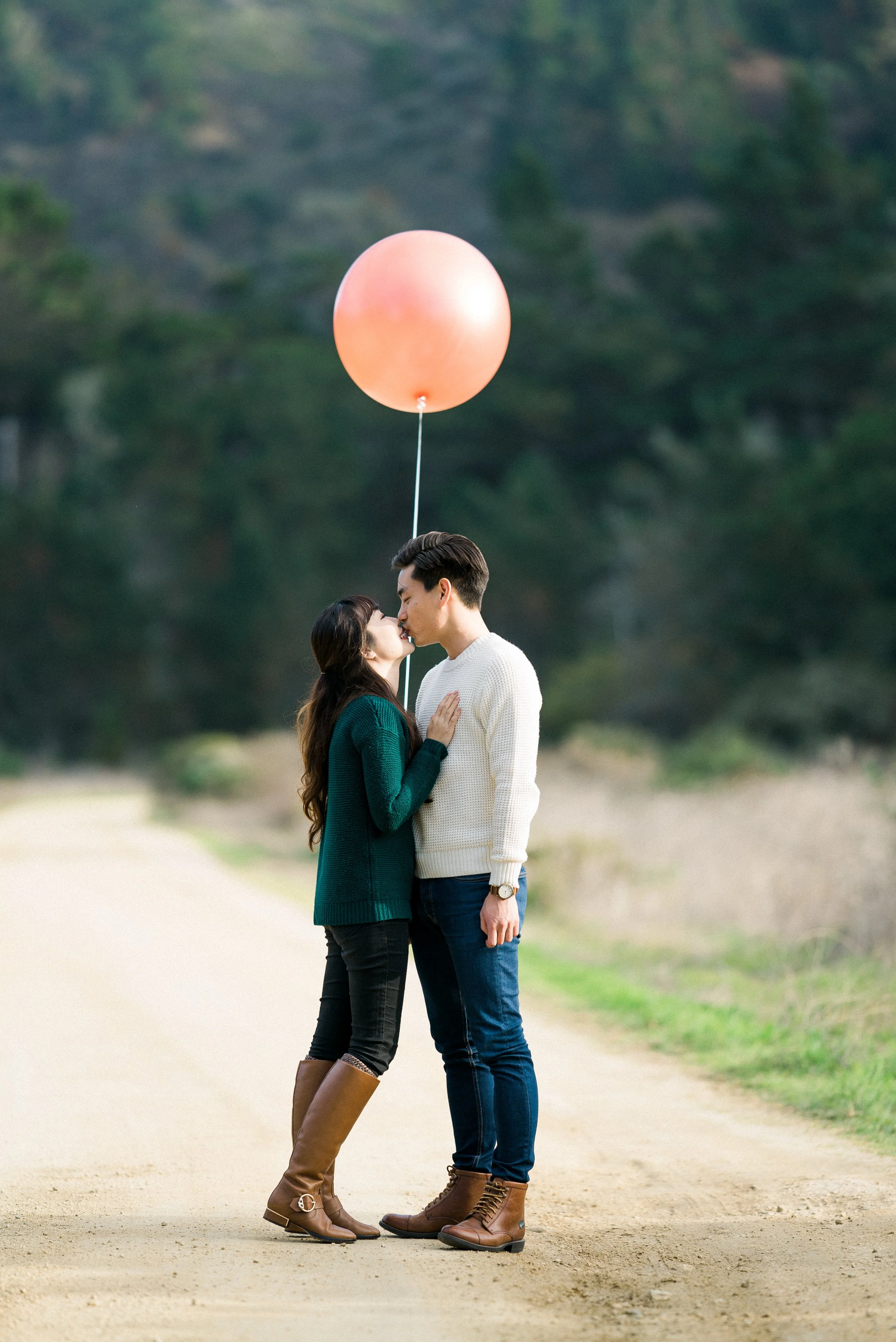 A couple kissing on a dirt path while holding a large peach-colored balloon, with a lush green forest background.