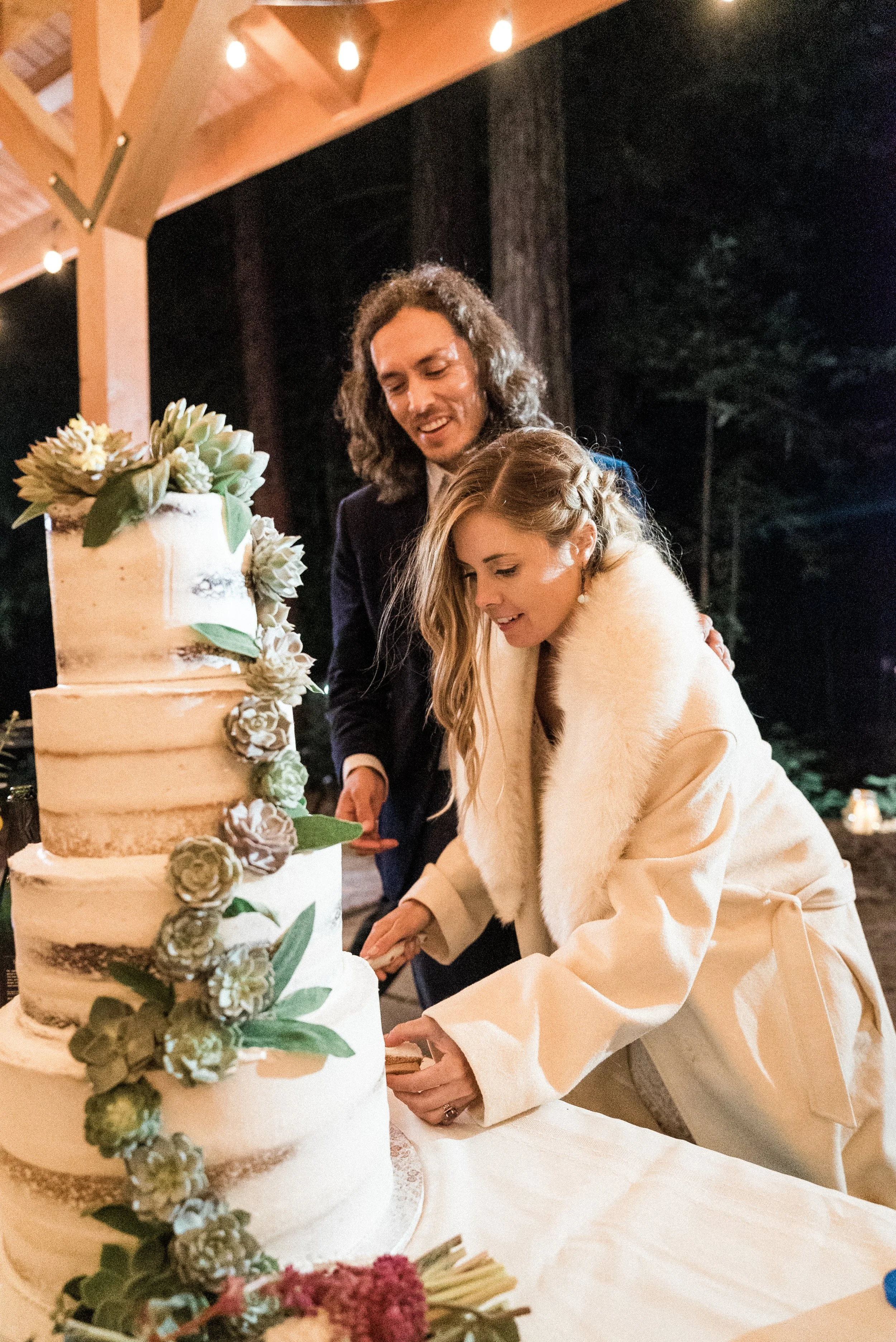 Bride wearing a white coat with a fur collar and groom with long wavy hair cutting a multi-tiered rustic wedding cake garnished with succulents during an outdoor evening reception.