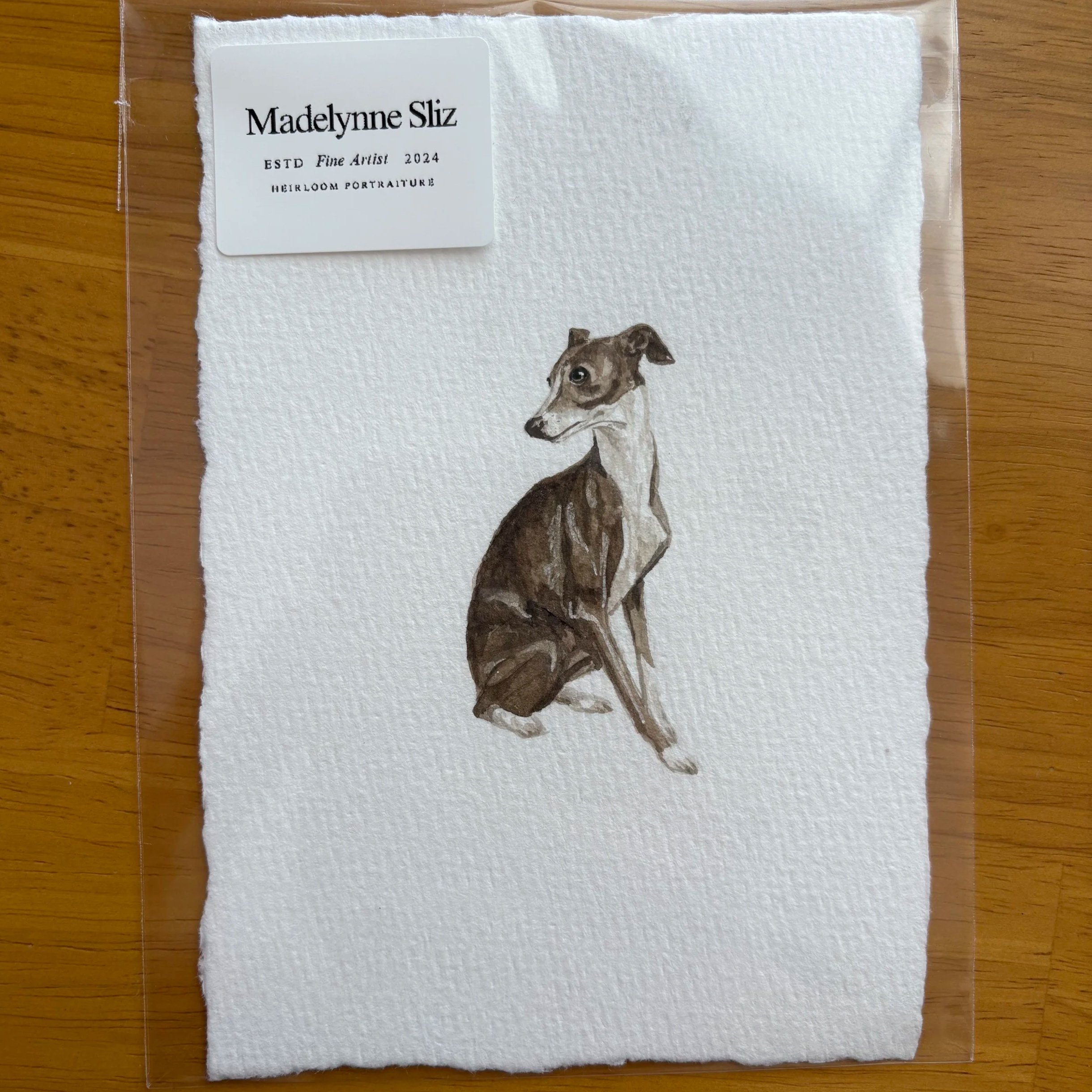 A watercolor portrait of a dog on textured white paper, with a label reading Madelynne Sliz, ESTD Fine Artist 2024, Heirloom Portraiture, on a wooden surface.