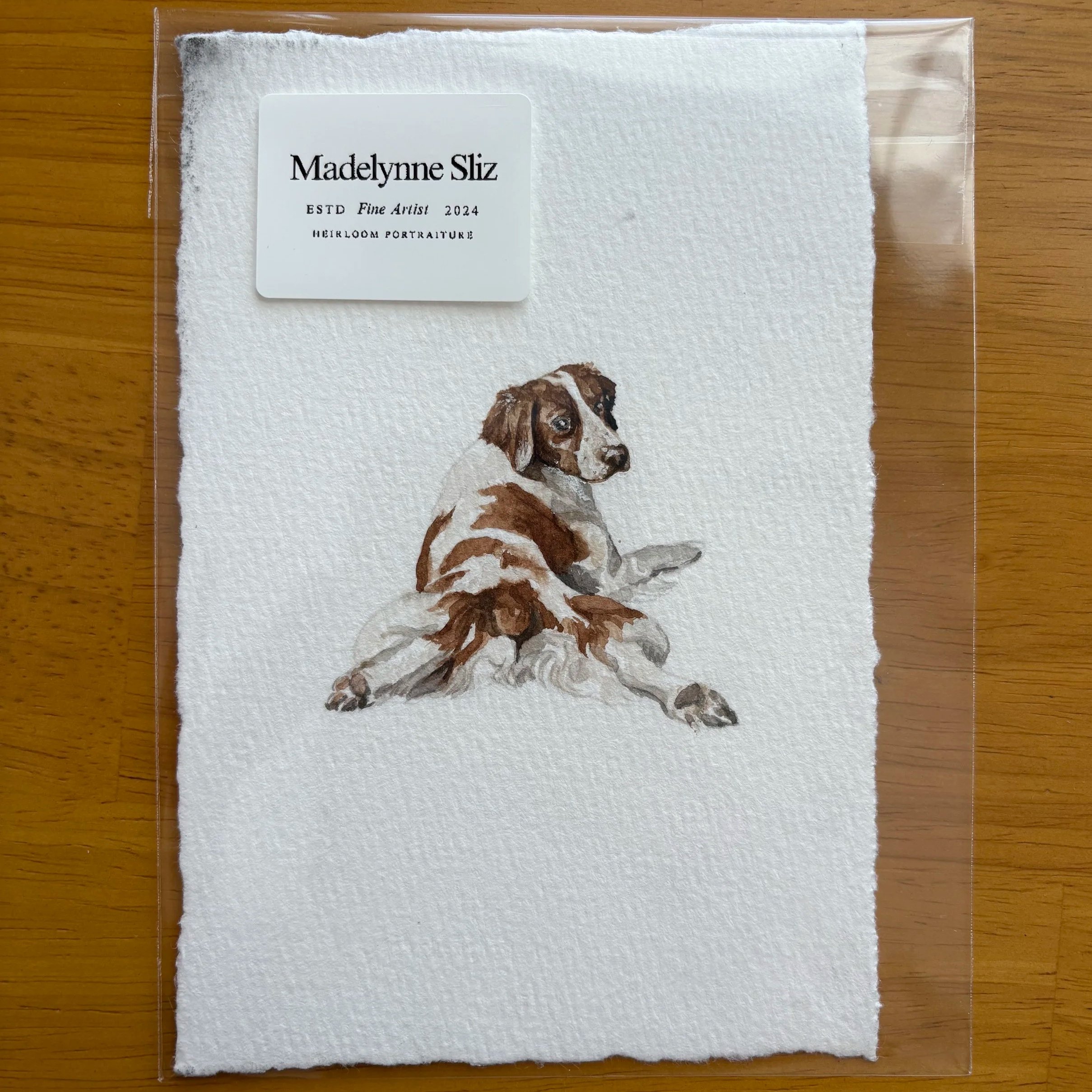 A piece of watercolor paper with a portrait of a dog, featuring a white background with brown spots, a brown patch over one eye, and floppy ears, placed on a wooden surface.
