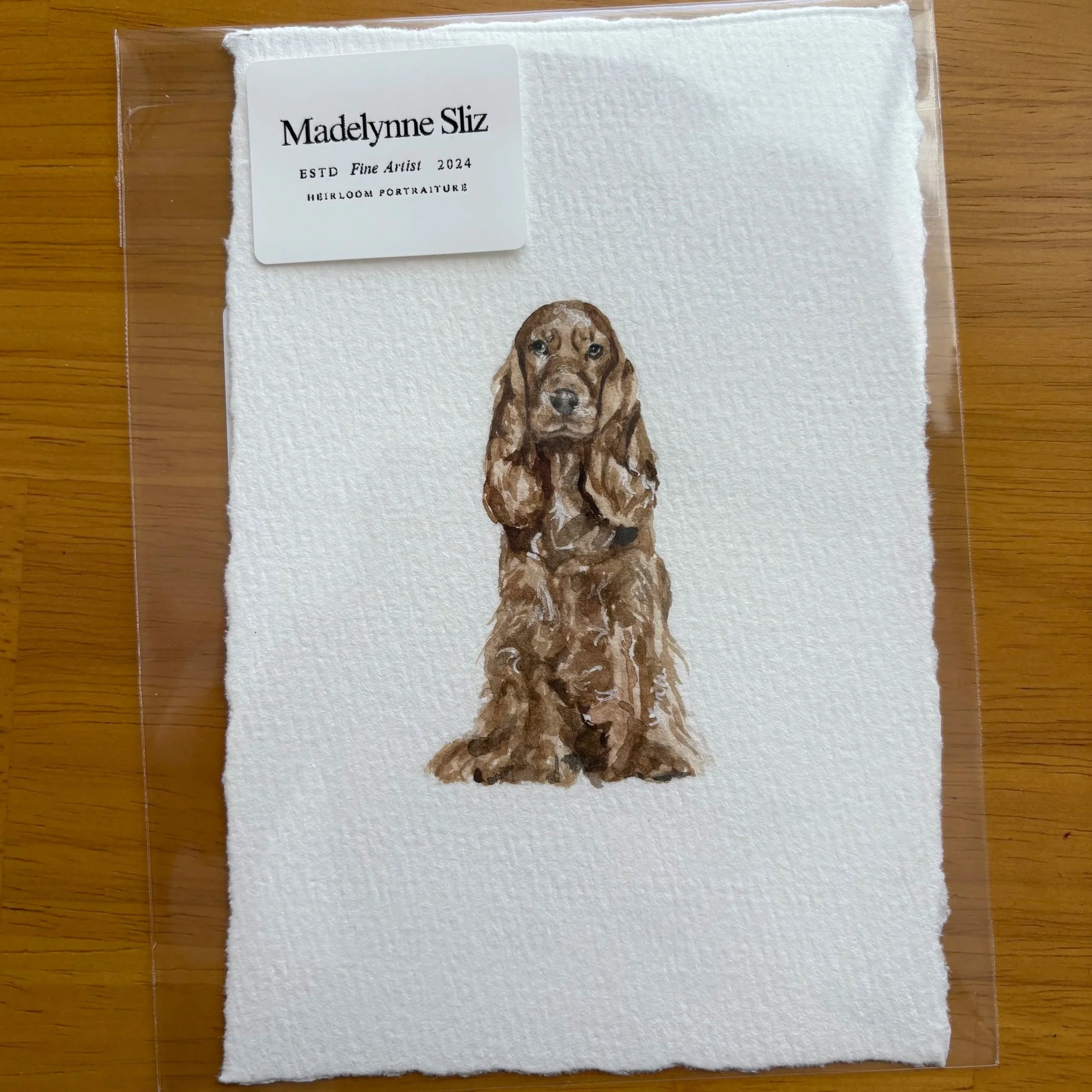 A towel with an illustration of a brown dog and a white sticker with black text that reads 'Madelynne Sliz ESTD Fine Artist 2024 HEIRLOOM PORTRAITURE' on a wooden surface.