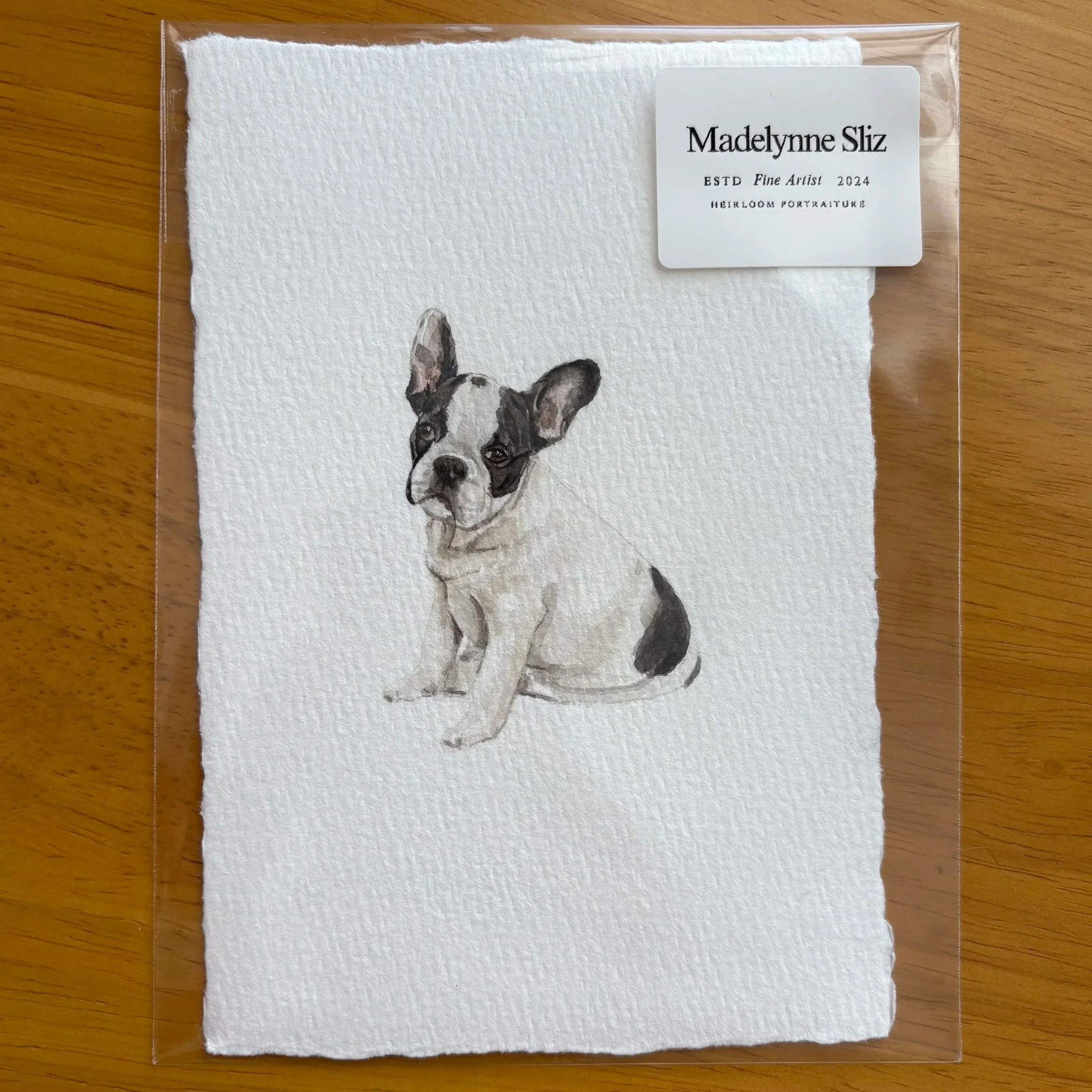 A watercolor portrait of a dog on textured white paper, inside a clear plastic sleeve with a white label reading 'Madelyne Sliz ESTD Fine Artist 2024 MEIRLOOM PORTRAITURE' on a wooden surface.