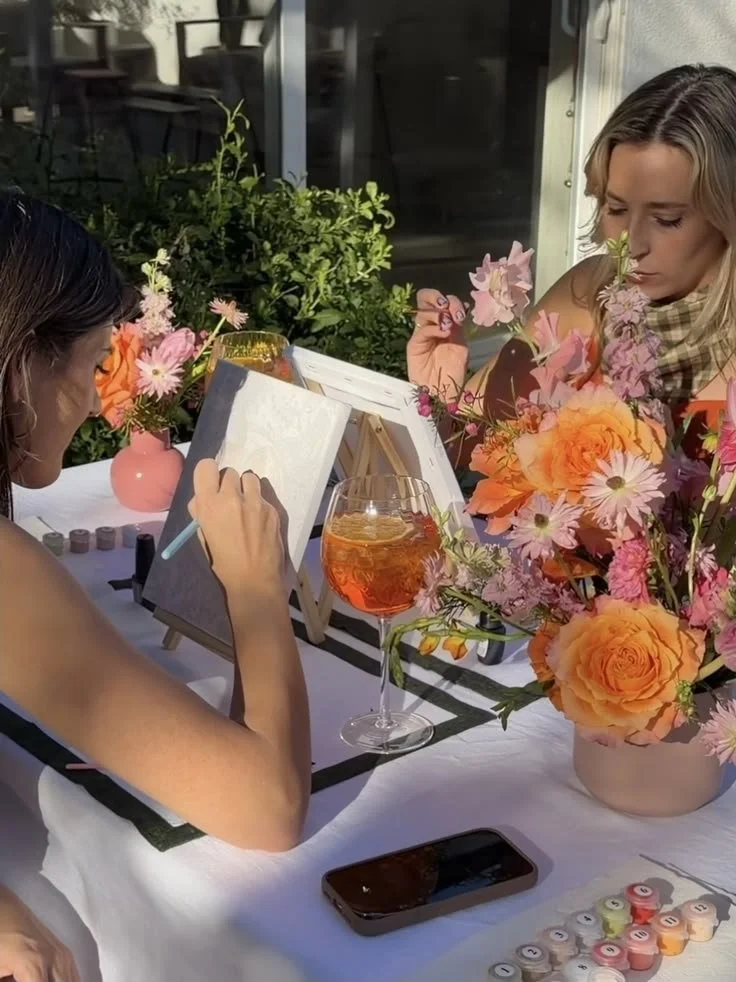 Two women sitting at a table outdoors, painting on canvases. The table is decorated with pink and orange flowers in vases, and there is a glass of orange-colored drink. A smartphone is on the table.