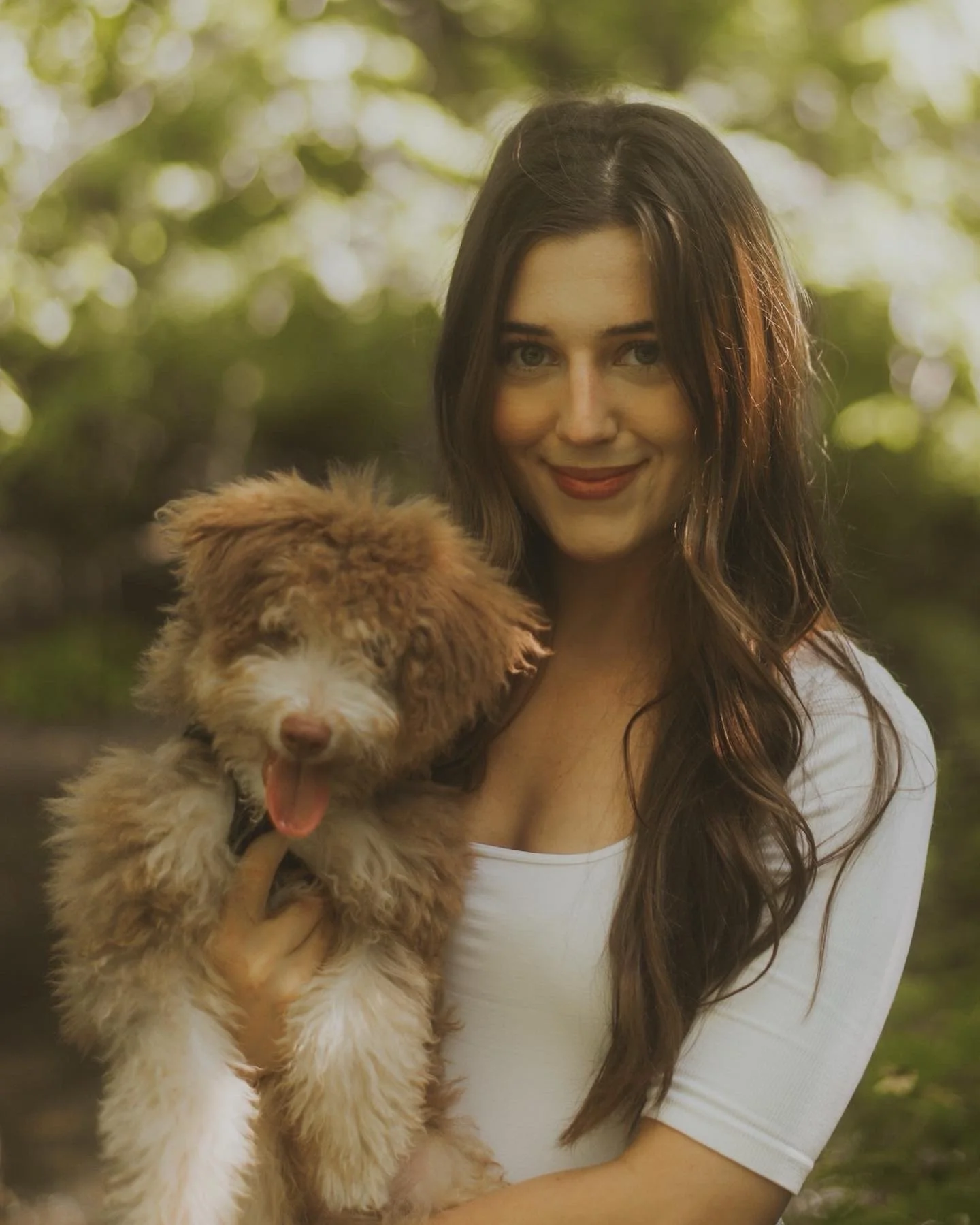 A woman with long brown hair smiling and holding a fluffy brown and white puppy with its tongue sticking out outdoors in a green, leafy area.