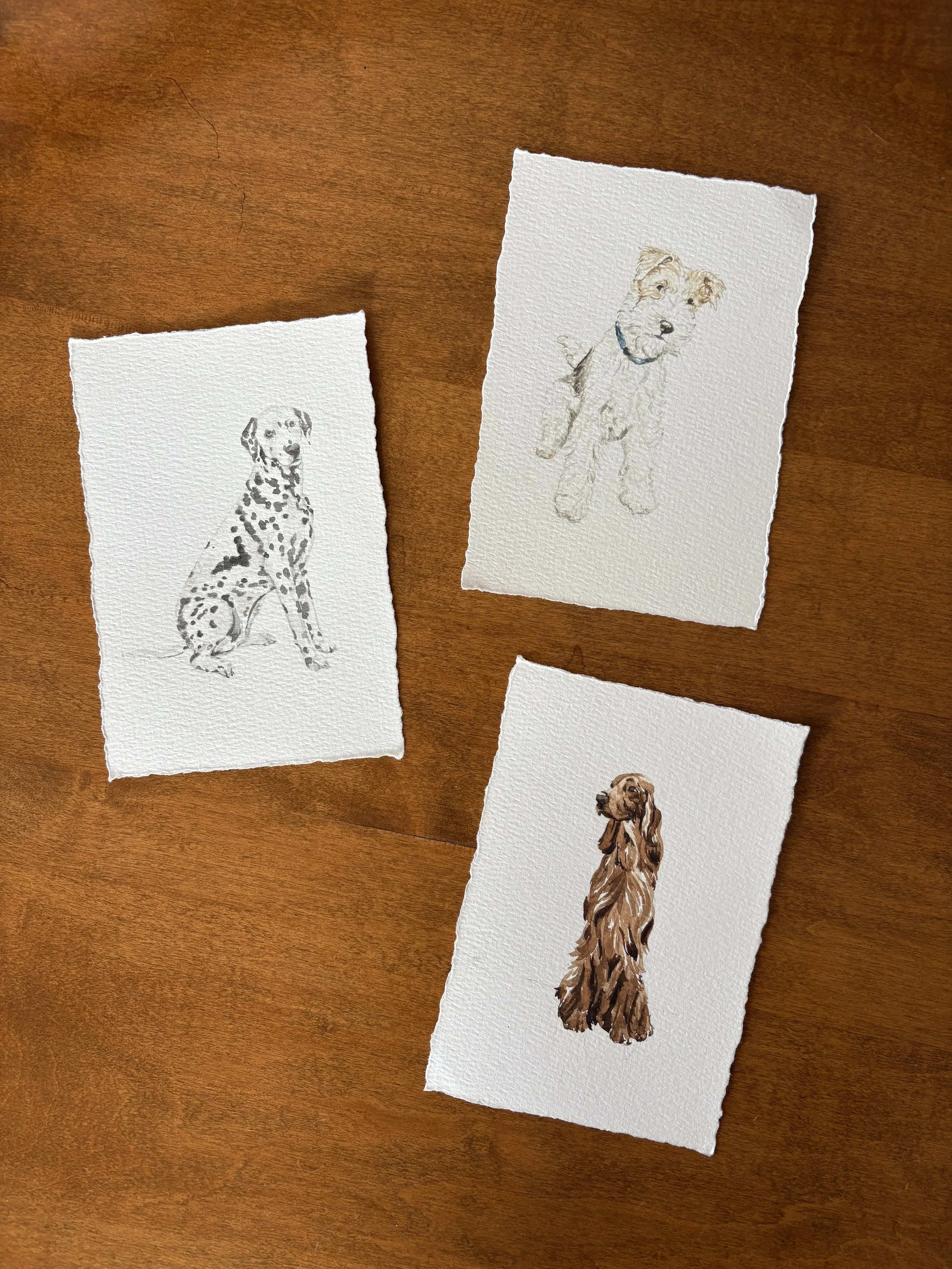 Watercolor paintings of three different dogs on textured paper, placed on a wooden surface.