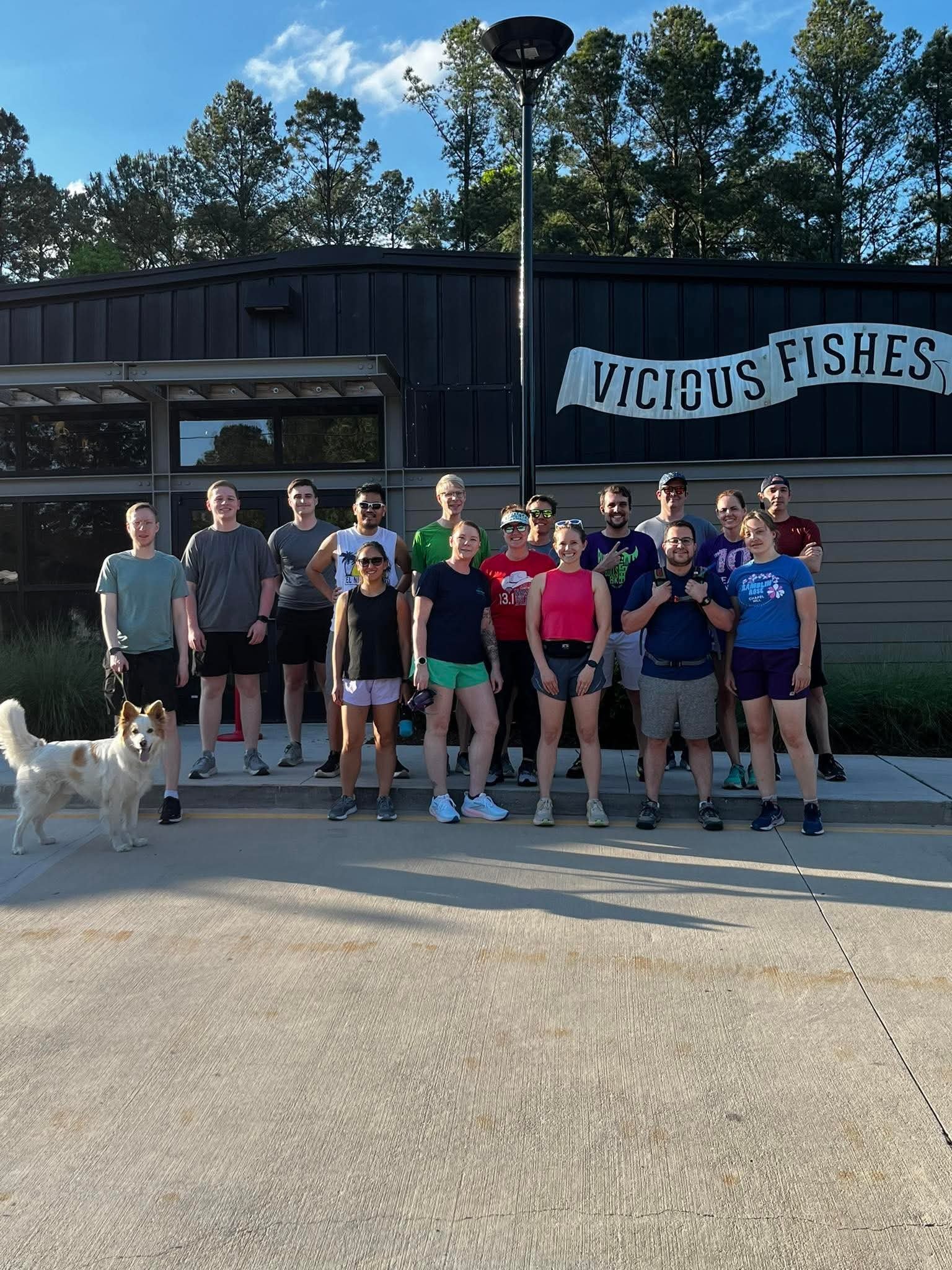 Group of people standing outside in front of a building with a sign that says "Vicious Fishes." There is a dog in the foreground on the left, and the group appears to be happy and smiling. Tall trees and a partly cloudy sky are in the background.