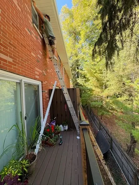 Person on a ladder fixing or inspecting the roof of a brick house from an outdoor deck, with potted plants and gardening tools nearby, and trees in the background.