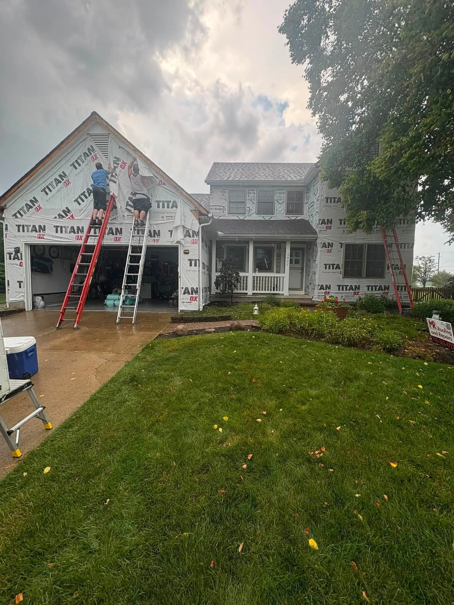 A house under construction with weather-resistant house wrap, two workers on ladders installing more wrap around the exterior, a driveway with some tools and construction materials, a partly cloudy sky, and green lawn with plants in the yard.