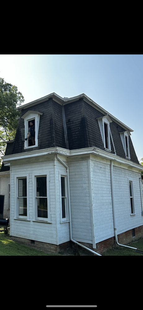 Side view of a two-story house with white siding and black dormer windows on the roof, a person peeking out of one of the dormer windows, green trees, and a clear blue sky.