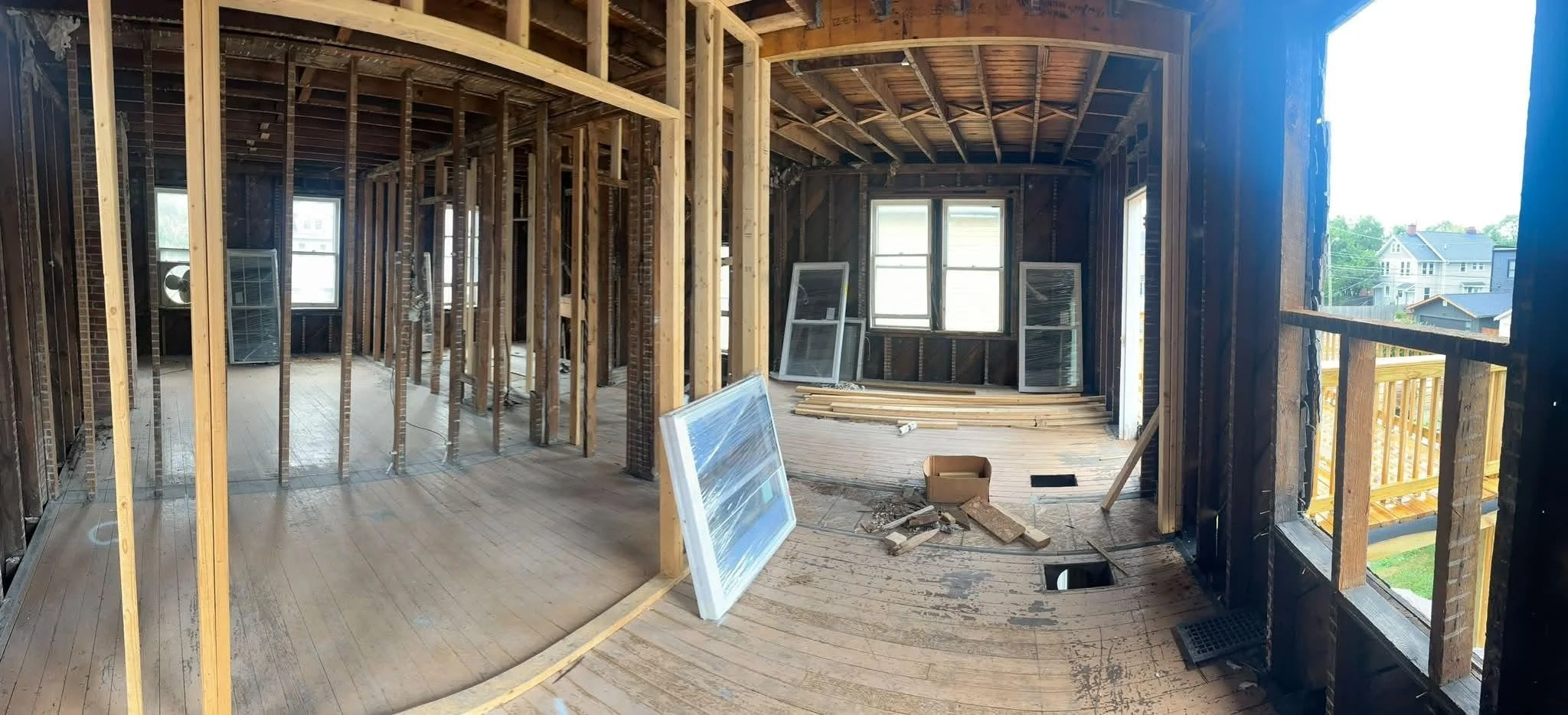 Interior of a house under construction with exposed wooden framing, windows, and kitchen area, showing unfinished walls and construction materials.