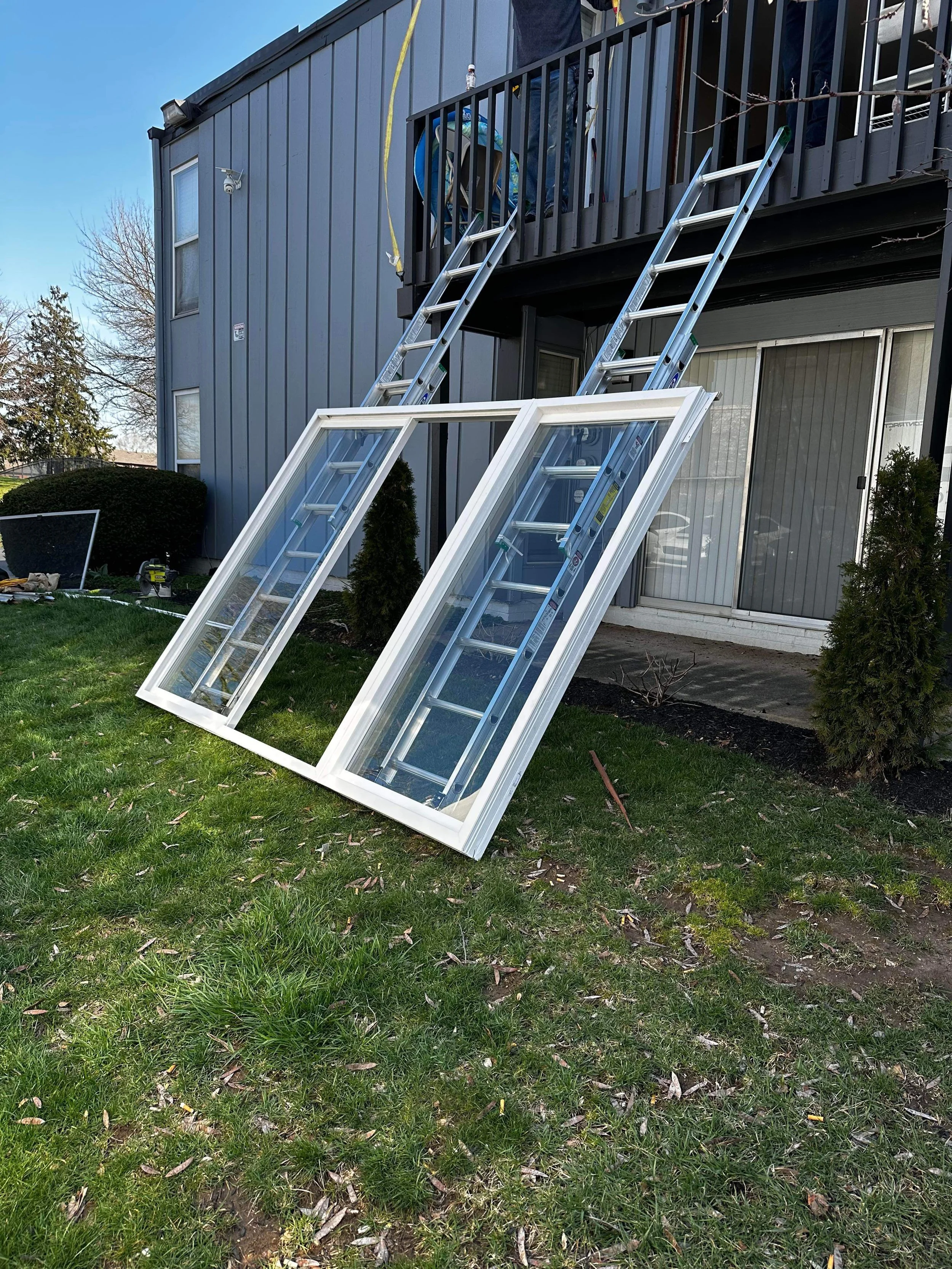 Two aluminum ladders leaning against a deck railing, with two glass windows on the ground level. A small grassy yard with bushes and a few tools or objects on the ground.