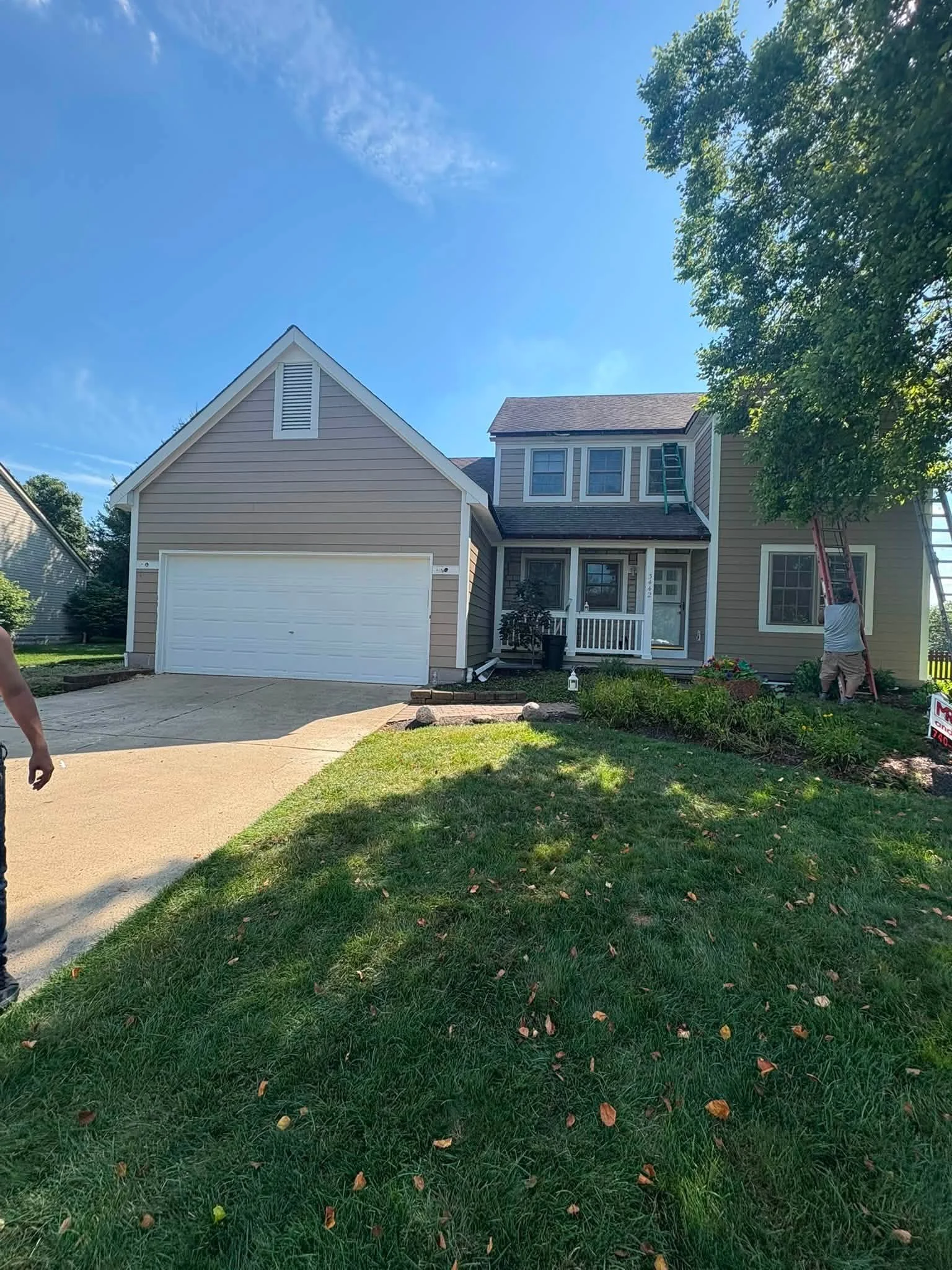 A two-story house with beige siding, a white garage door, and a front porch with a small railing. Several trees surround the house, and a person is seen standing on a ladder onto the roof. The lawn is green with fallen leaves, and the sky is mostly c