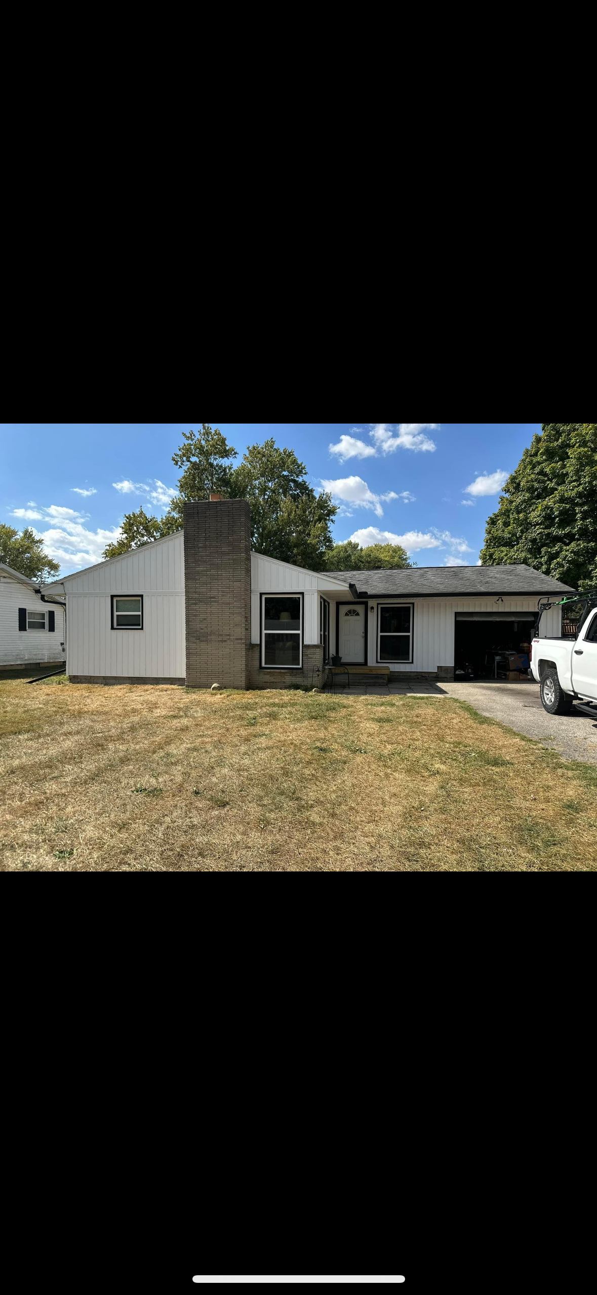 A single-story house with white siding, black shutters, and a brick chimney, surrounded by a yard with dry grass, beside a driveway with a white pickup truck parked outside the garage, under a partly cloudy sky with green trees in the background.
