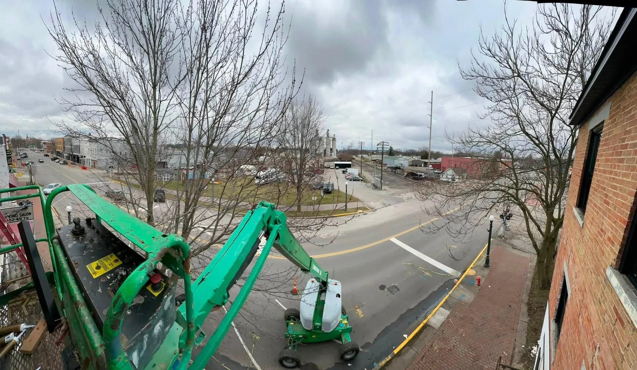 View from a building window shows leafless trees, a street with parked cars, and a few commercial buildings under cloudy sky. There is construction equipment visible in the foreground.