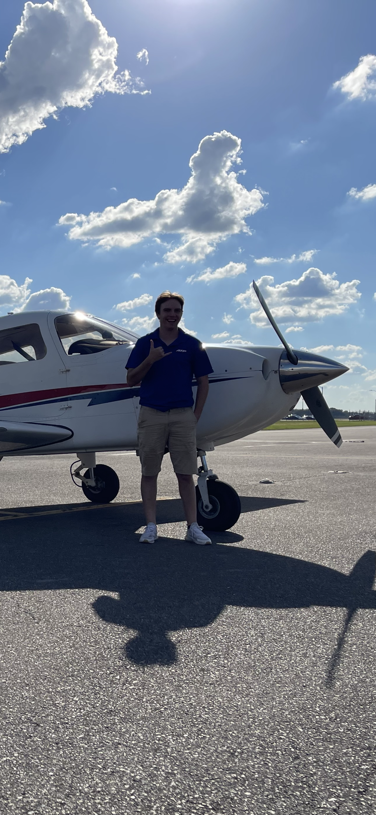 Young man standing in front of a small aircraft on an airport tarmac, making a shaka sign with his right hand, with a partly cloudy blue sky overhead.