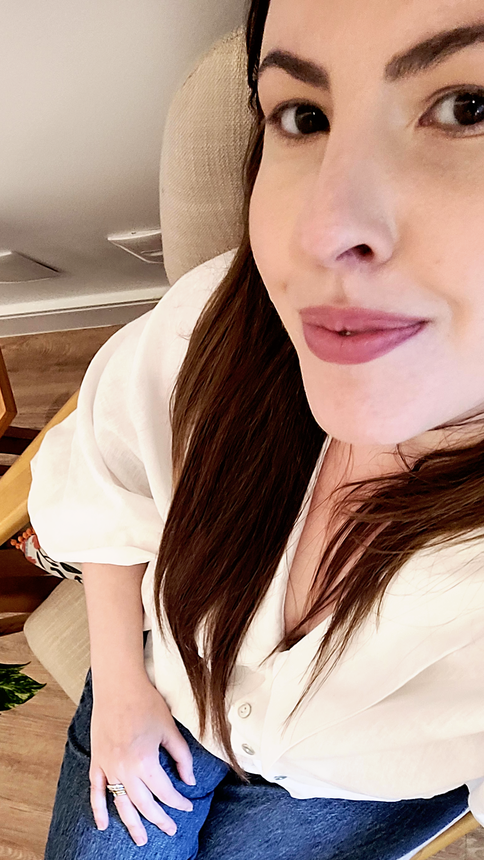 Mulher com cabelos longos, pele clara, usando blusa branca, sentado em uma cadeira de couro e sorrindo levemente, em ambiente interno.