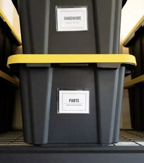 A black plastic storage bin with a yellow handle, labeled with two signs: 'Hardware' on the top and 'Parts' on the front, on a wire shelf.