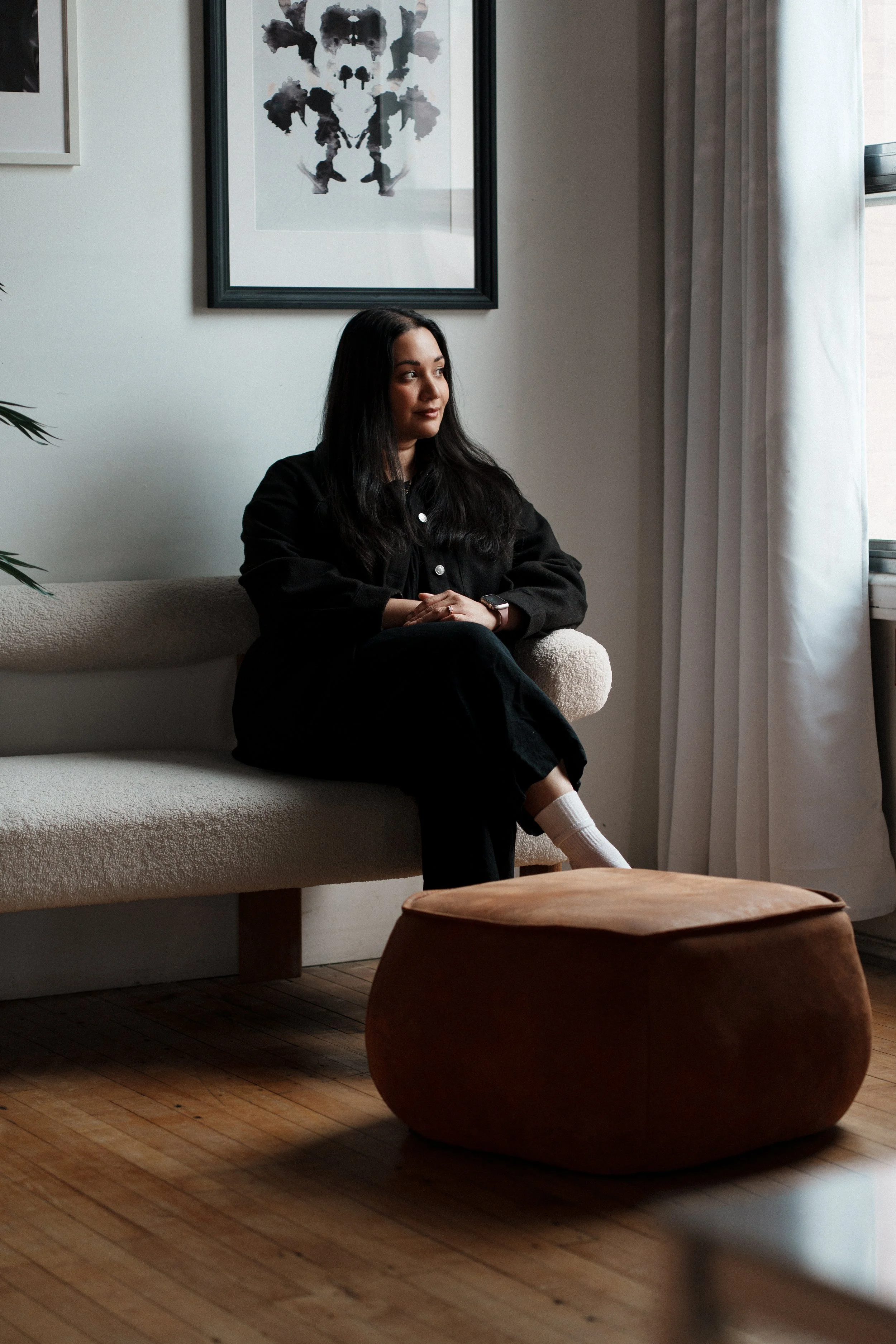 A woman with long dark hair, dressed in black, sits on a beige sofa in a room with minimalist decor, a framed black and white animal-themed artwork on the wall, and white curtains on a window to the right.