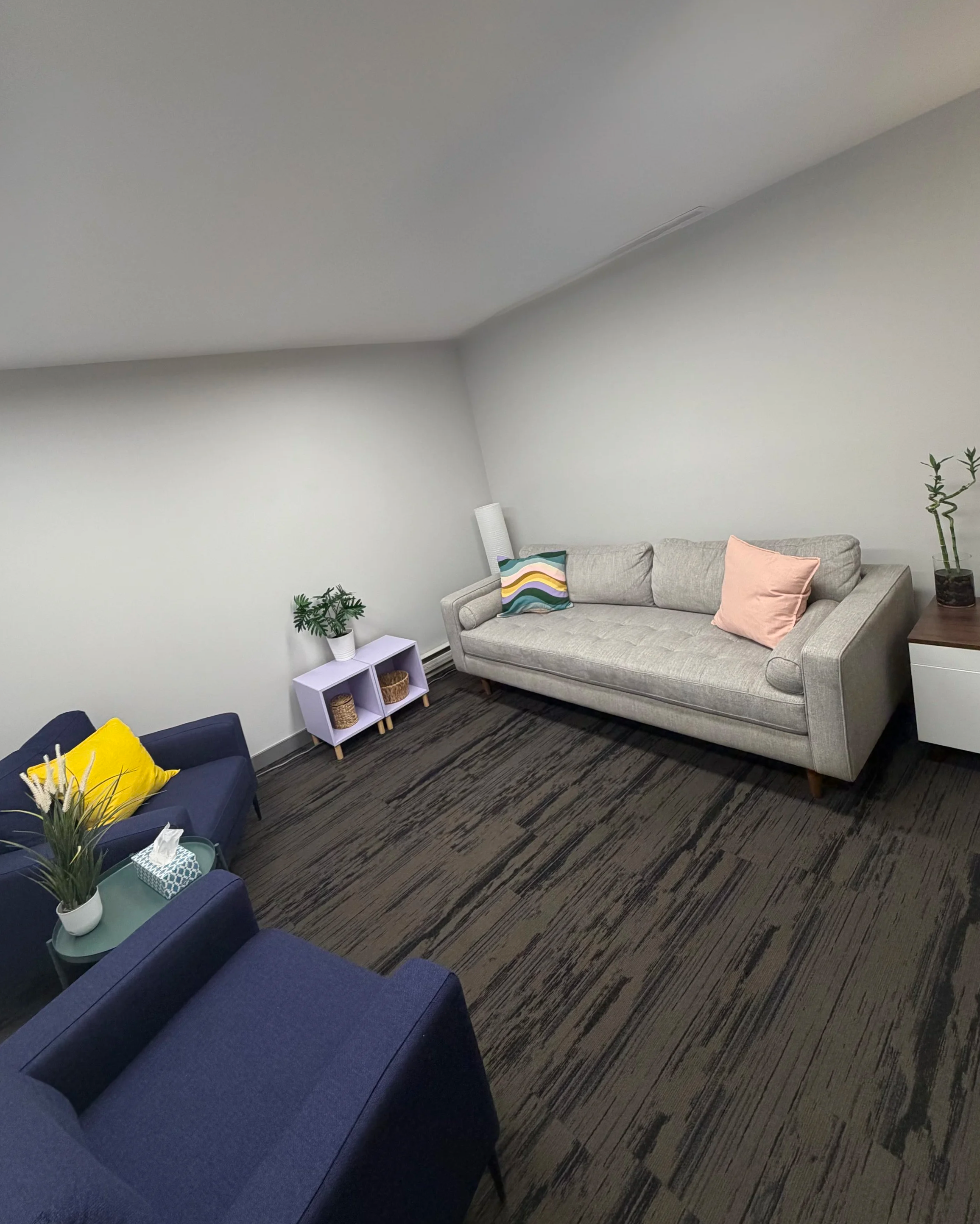 A living room with a gray couch decorated with a colorful zigzag pattern pillow and a pink pillow. A white side table with a plant and decorative containers is beside the couch. A blue armchair with a yellow pillow is in the foreground. The floor has a striped dark-colored carpet.