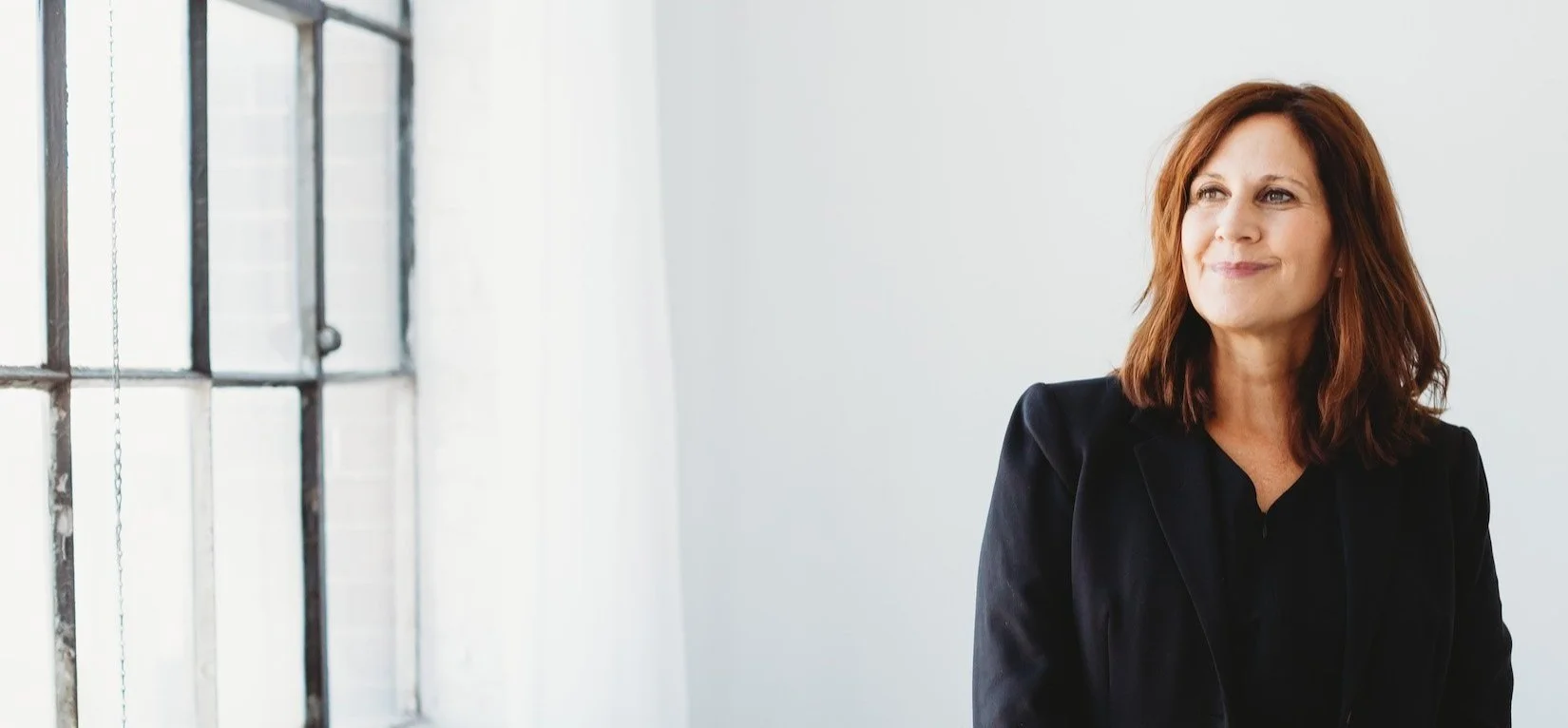 A woman with shoulder-length auburn hair and a black blazer standing near a large window with a white wall in the background.