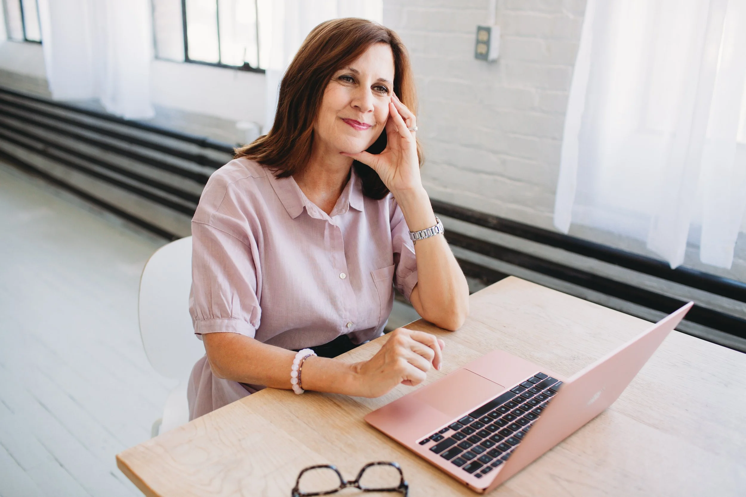 A woman with shoulder-length brown hair wearing a light pink shirt sits at a wooden table with a pink laptop, resting her chin on her hand and smiling, in a bright room with large windows and a white brick wall.