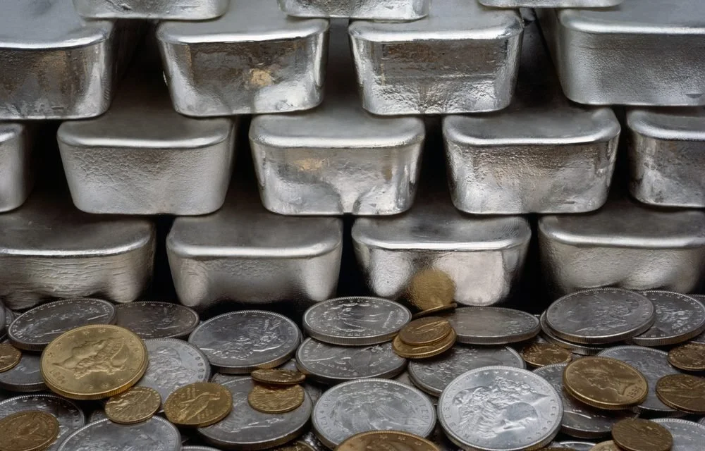 Stacks of 100oz t silver bars and scattered U.S. coins, Dollars, Half Dollars, Quarters, Dimes, Nickels, and Cents.